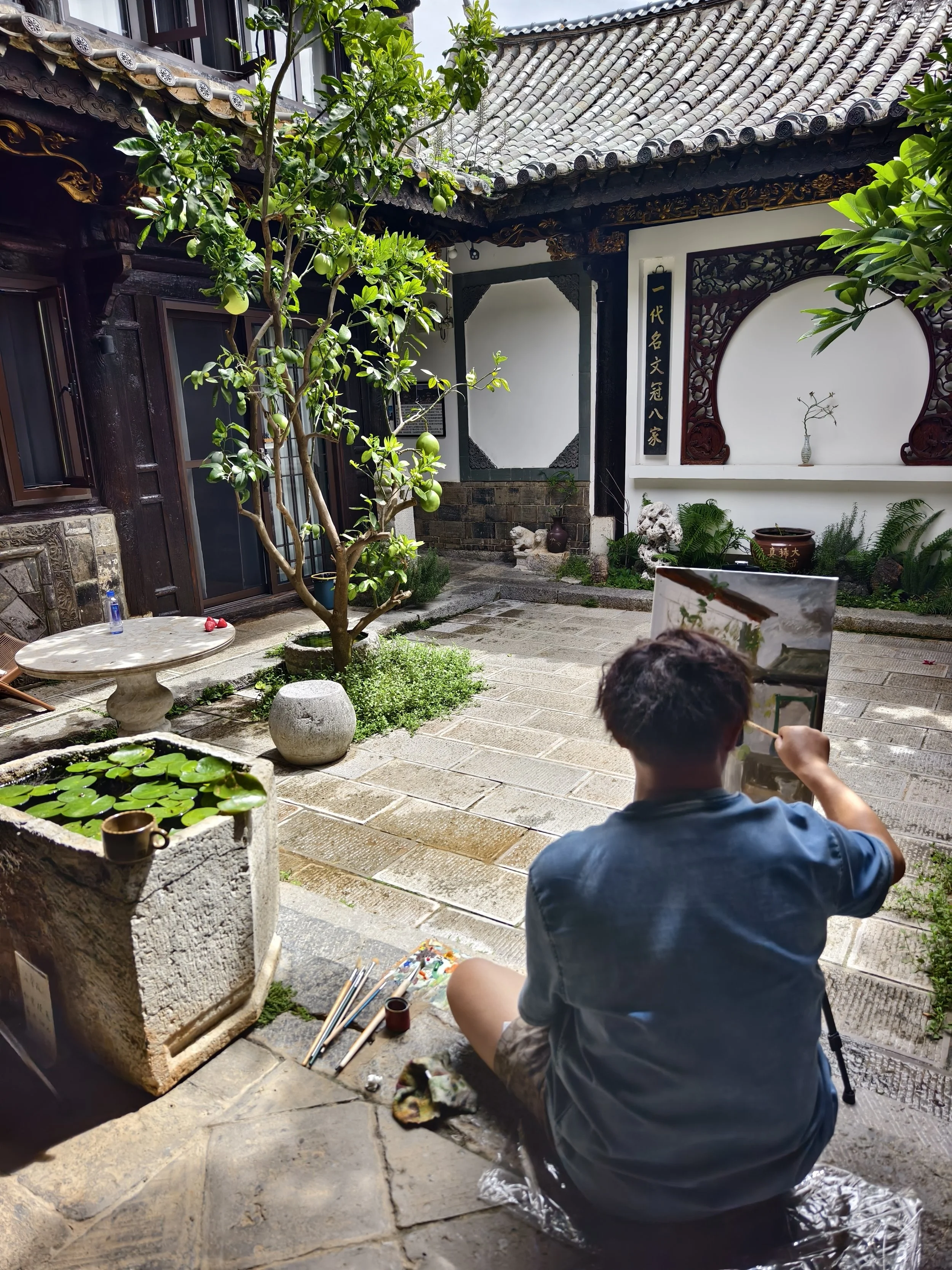 A seated figure beside a courtyard pond, surrounded by plants and tiled-roof buildings.