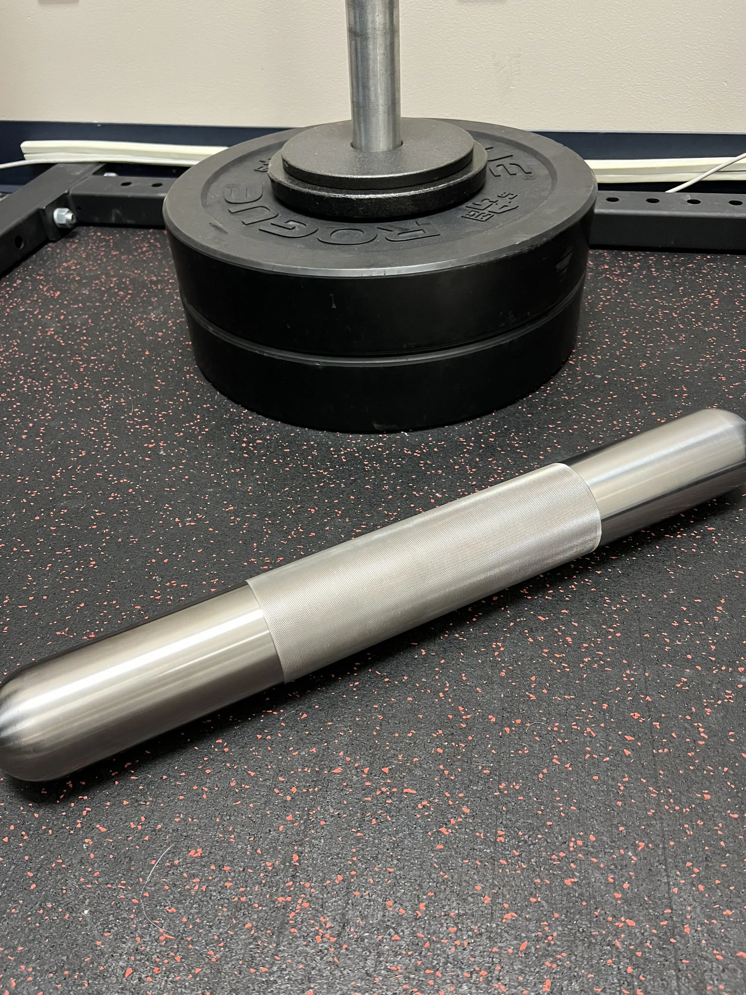 A barbell with weights resting on a black and red speckled gym floor, a weight plate with the brand 'Rogue' on top, and a silver textured handle lying on the floor.