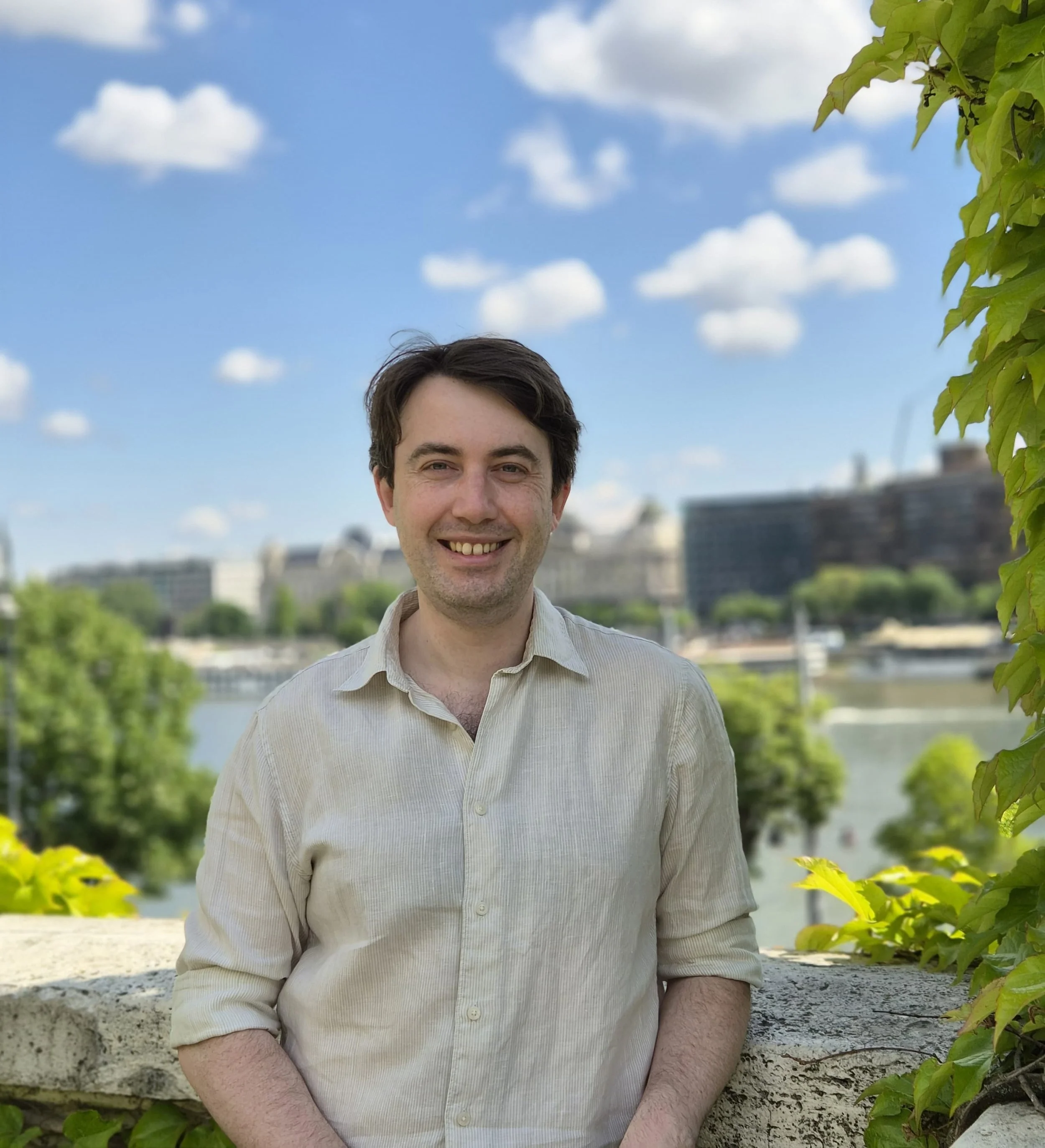 A smiling man with dark hair, wearing a light-colored shirt, standing outdoors on a sunny day with a bright blue sky and scattered clouds, with green leaves and trees in the background.