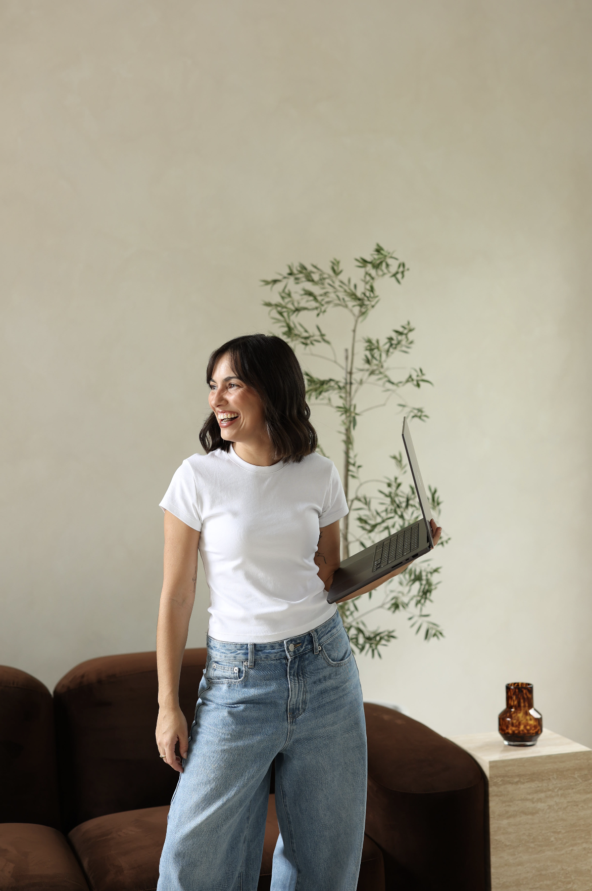 A woman with dark hair, wearing a white t-shirt and jeans, smiling and holding a laptop in a living room with a green plant and brown couch.