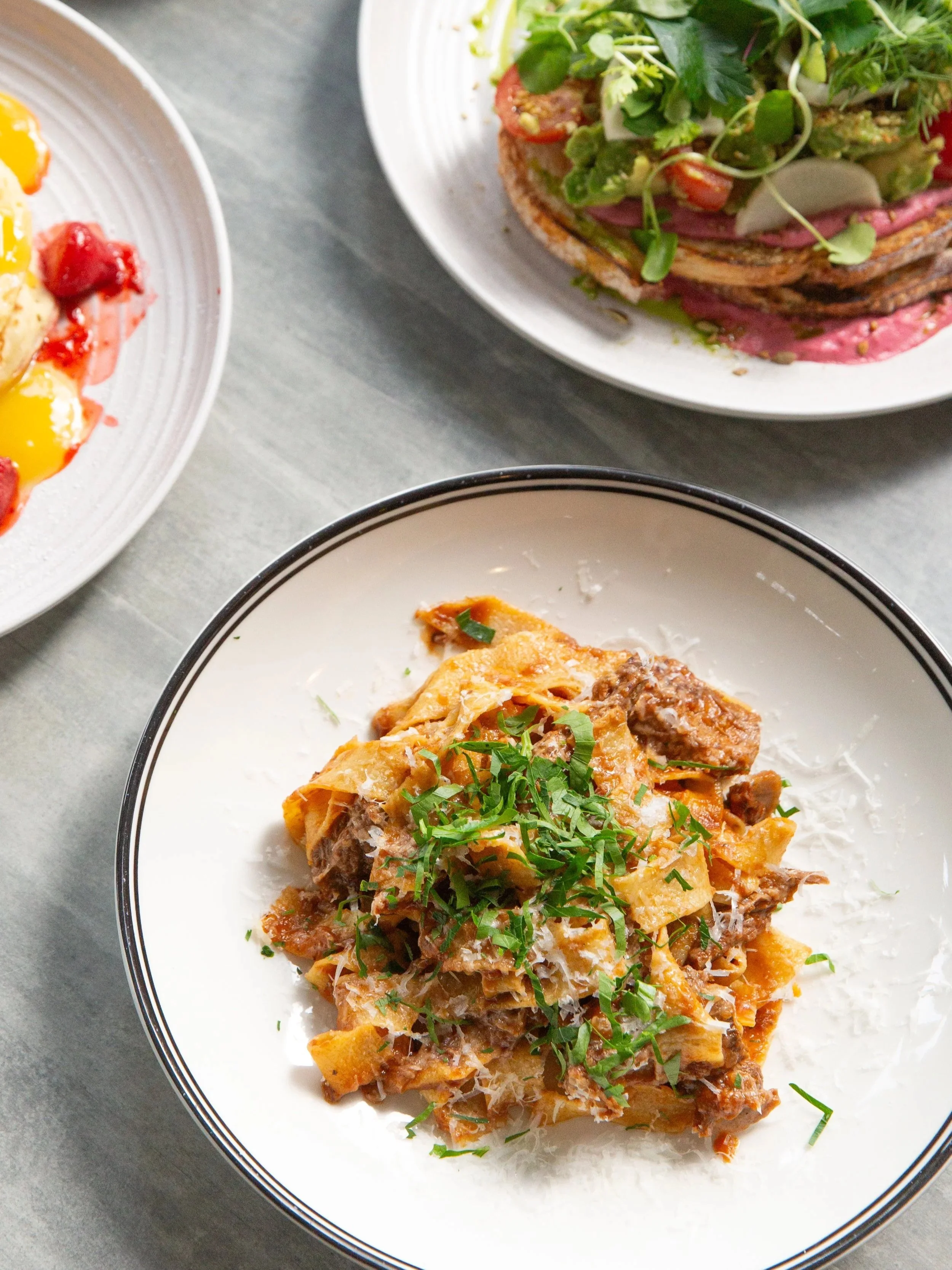 A plate of lasagna garnished with chopped herbs, with other dishes partially visible on adjacent plates, on a gray surface.