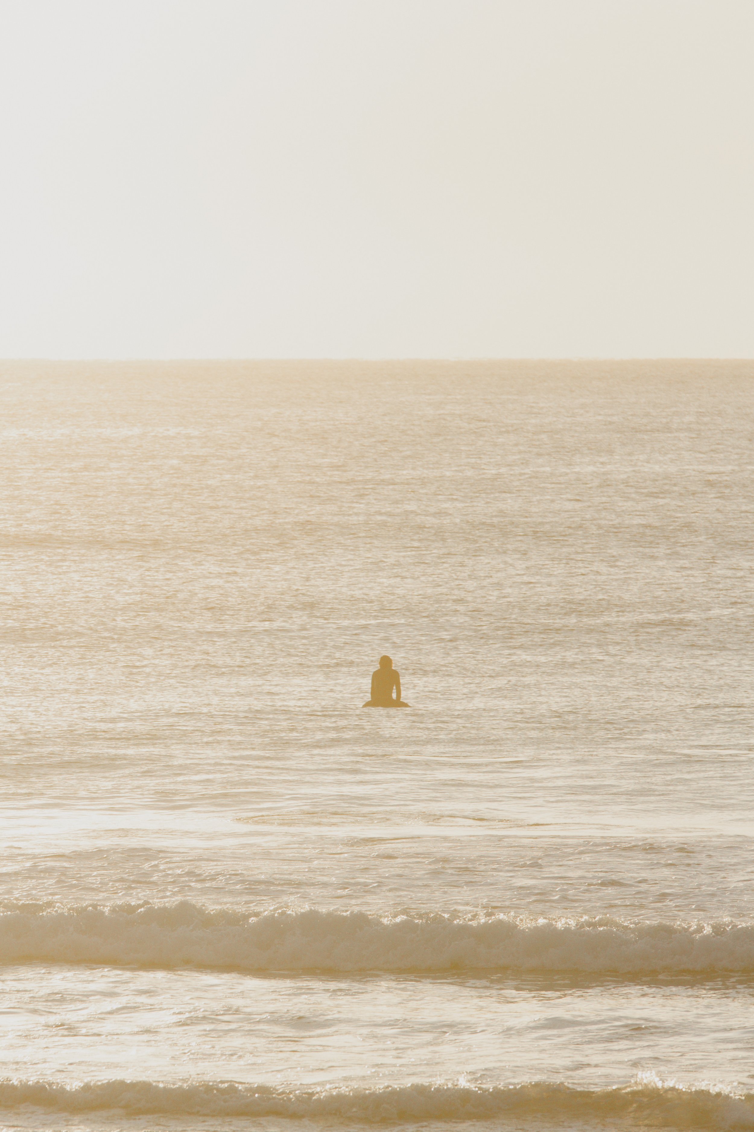 A person sitting alone on a beach, facing the ocean during sunset or sunrise, with calm waves and a slightly hazy sky.