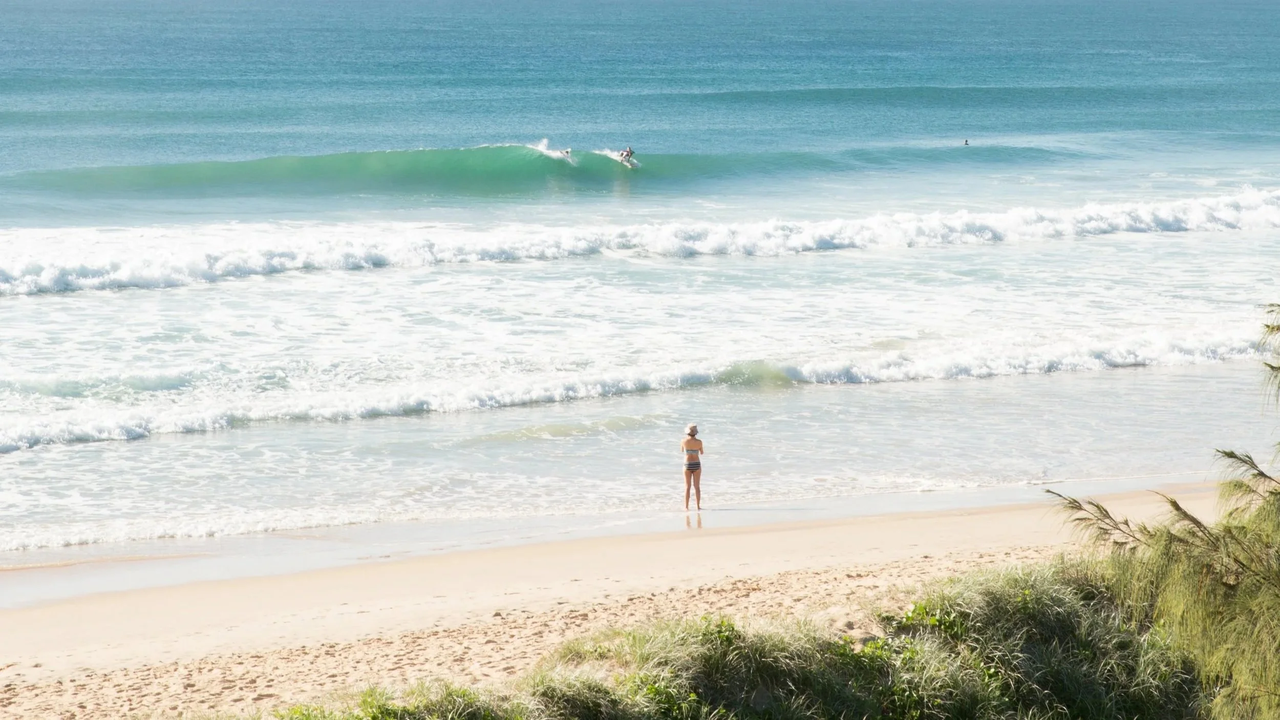 Woman standing on the sandy beach watching surfers ride the waves in the ocean.