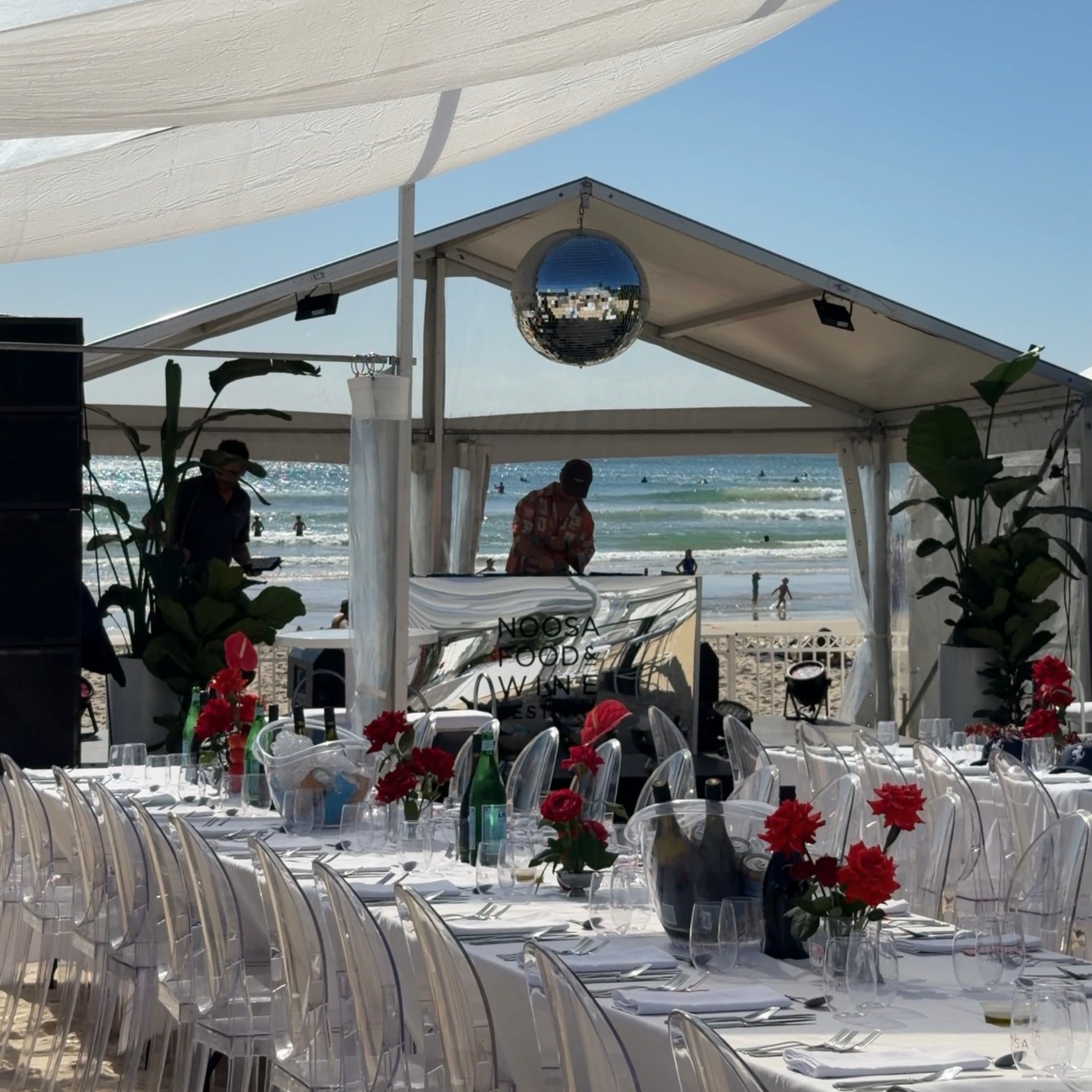 Beachside outdoor event setup with long dining tables covered with white tablecloths, decorated with red flowers and glassware, under a white canopy. There are clear acrylic chairs around the tables, a small stage or DJ booth in front, and the ocean with swimmers and people enjoying the water in the background.