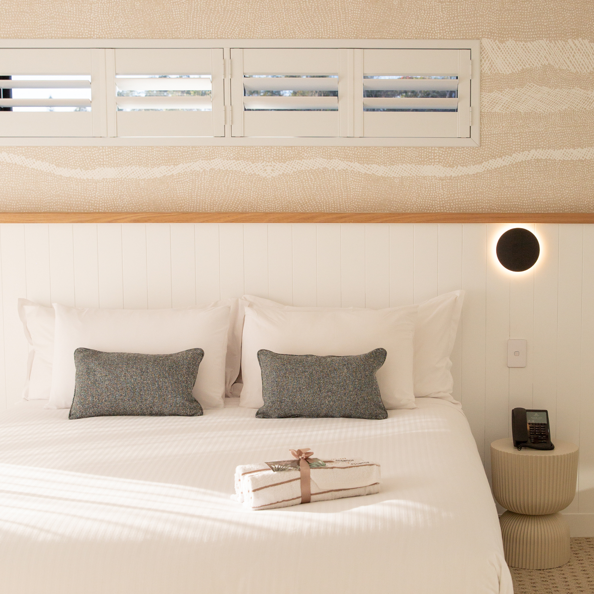 A neatly made hotel or bedroom with white bed linens and matching pillows. On the bed, there is a small, wrapped gift with a pink ribbon. The headboard area includes two decorative pillows and a bedside table with a telephone. Above the bed, there is a window with white privacy blinds, and a section of textured, patterned wallpaper or wall covering. The overall ambiance is clean and inviting.
