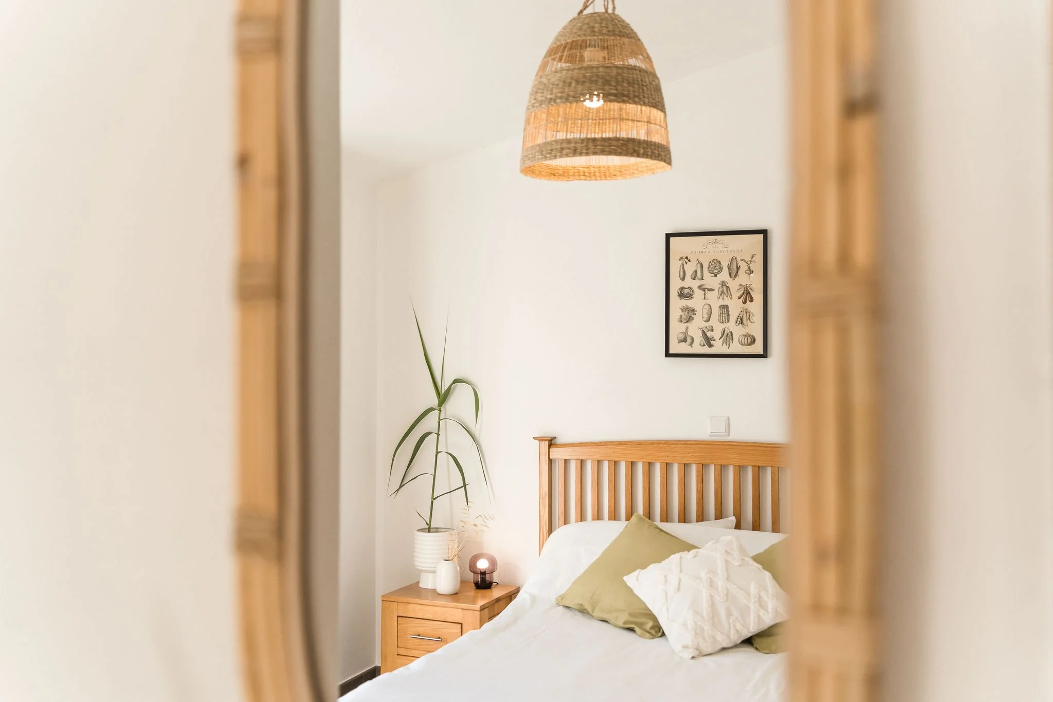 Ein Blick in ein Schlafzimmer, gesehen durch einen offenen Holzrahmen. Das Zimmer hat ein Bett mit weißen Bettwäsche und grünen Kissen, eine Nachttischlampe und eine Pflanze. An der Wand hängt ein Bilderrahmen mit Illustrationen von Gemüse und Frücht
