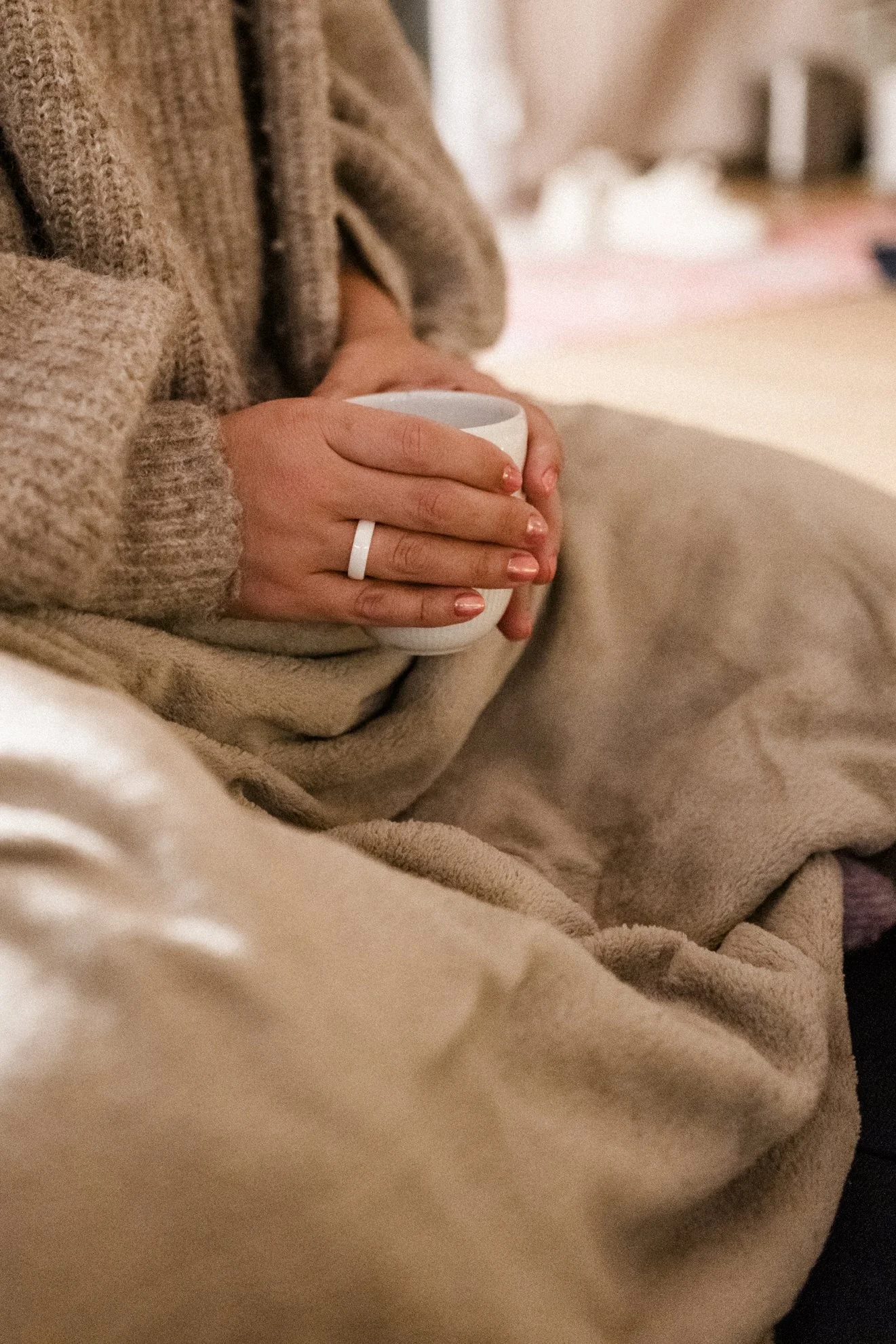 Person hält eine weiße Tasse mit beiden Händen, trägt eine braune Wolljacke und einen Ring am Finger, sitzt bequem in einem gemütlichen Raum.