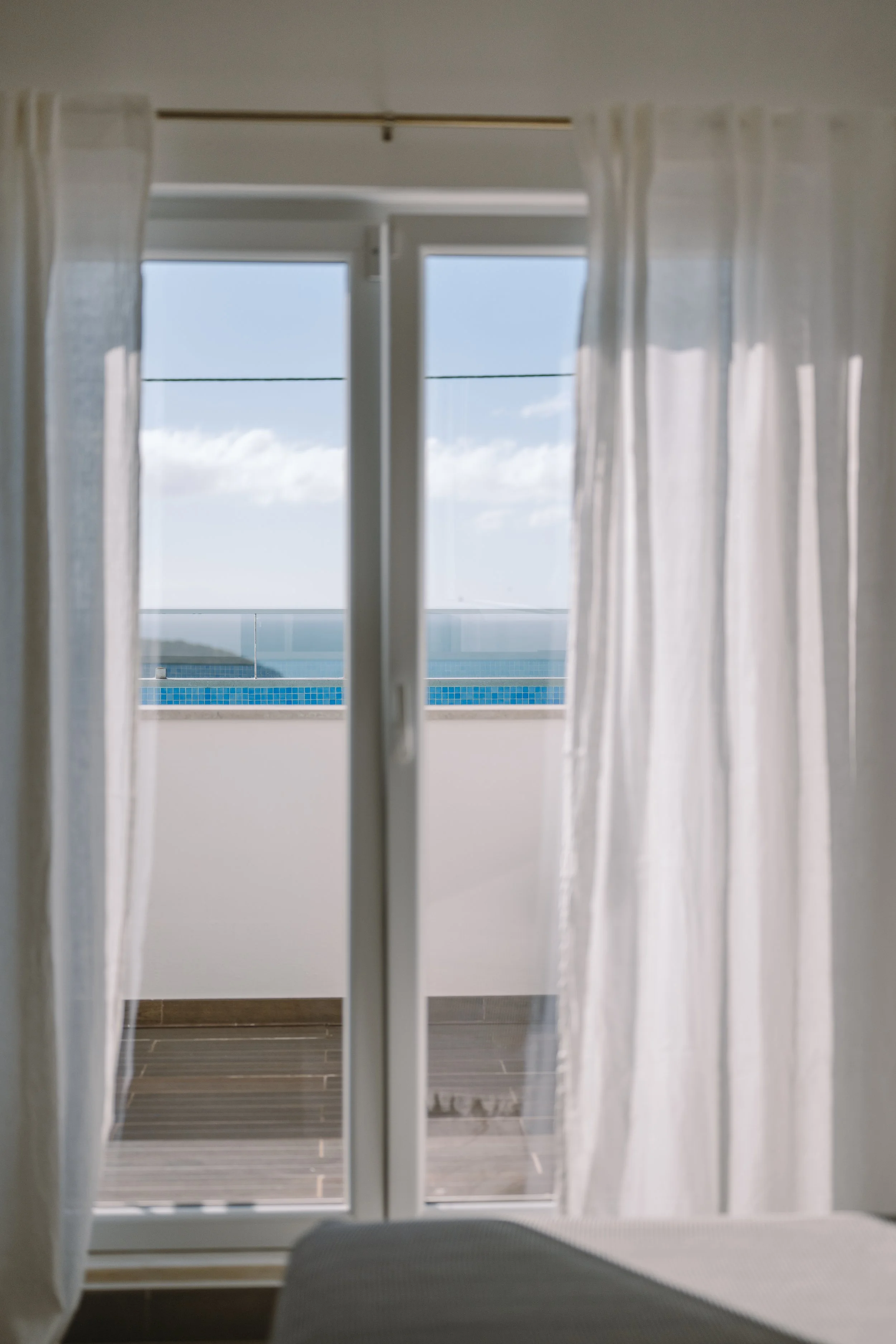 Blick durch eine geschlossene Glastür auf einen Balkon mit blauer Fliesenverkleidung, Blick auf den Himmel und die See im Hintergrund, weiße Vorhänge an den Seiten.