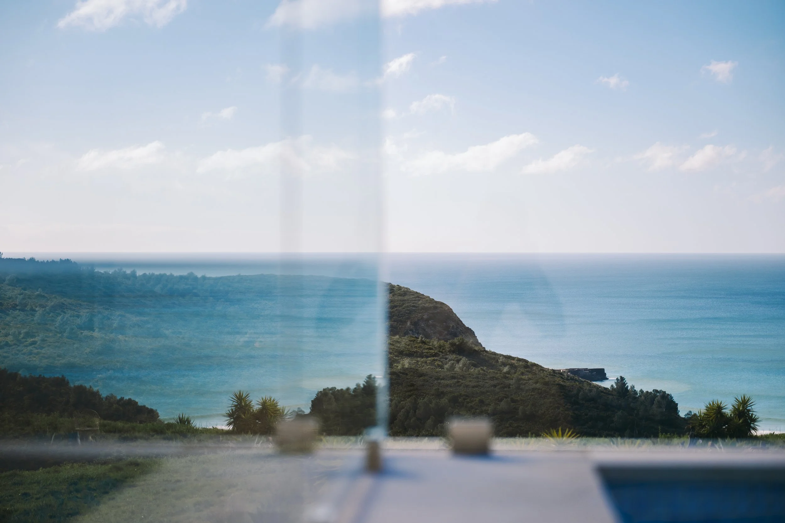 A scenic view of the ocean and coastline seen through a window with vertical blinds, with a lush green foreground and a partly cloudy sky.