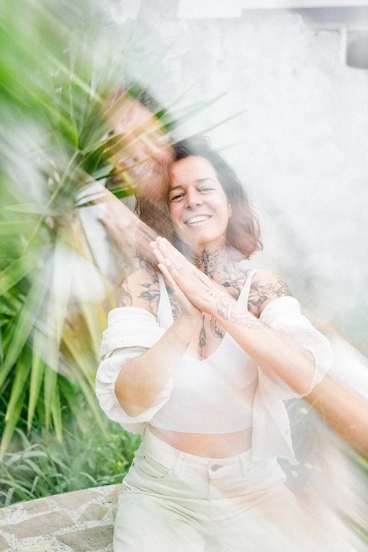 Eine lachende Frau mit Tattoos, die ihre Hände zusammenlegt, hinter einer Glasscheibe mit reflektierenden Lichtstreifen, umgeben von grünen Pflanzen.
