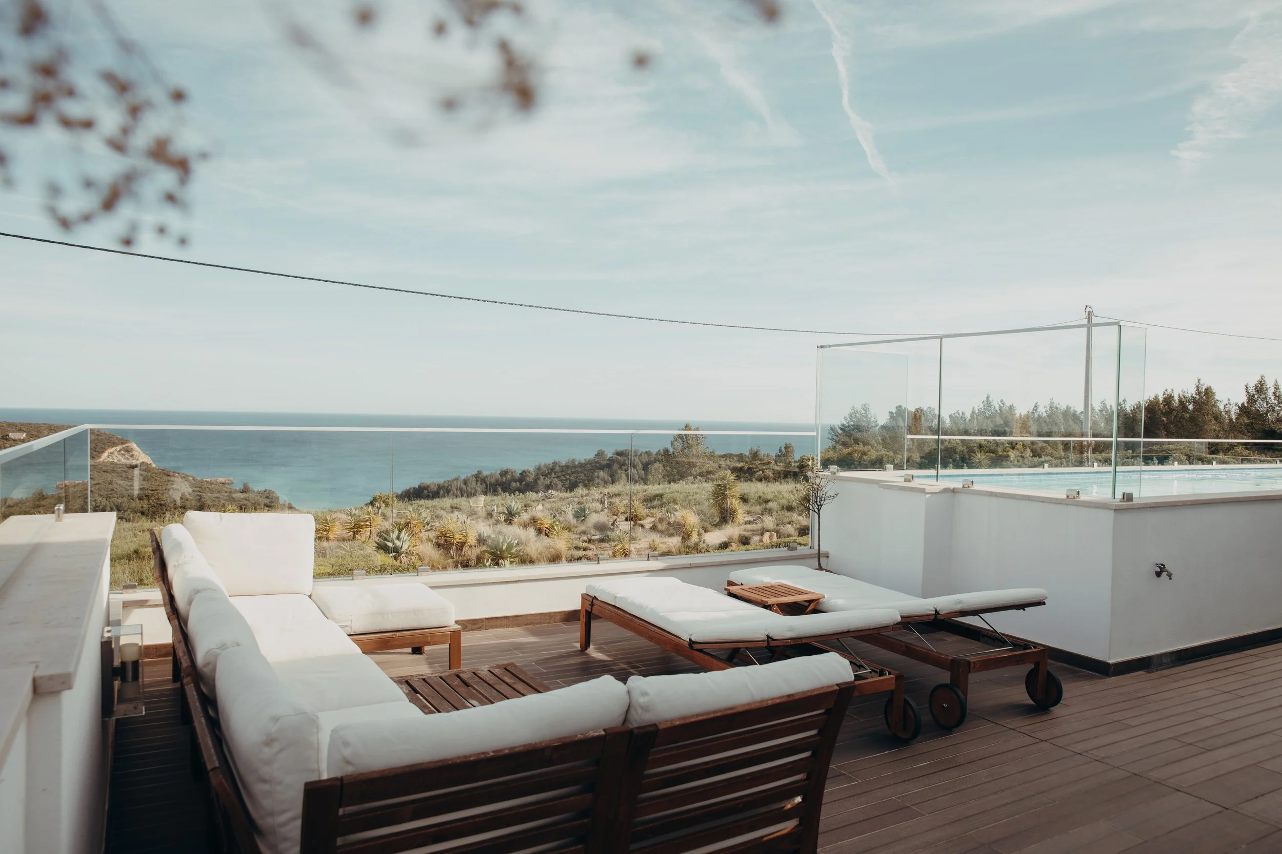 Ein Balkon mit weißen Sitzmöbeln, Blick auf das Meer und eine Landschaft mit Büschen und Bäumen im Hintergrund bei sonnigem Wetter.