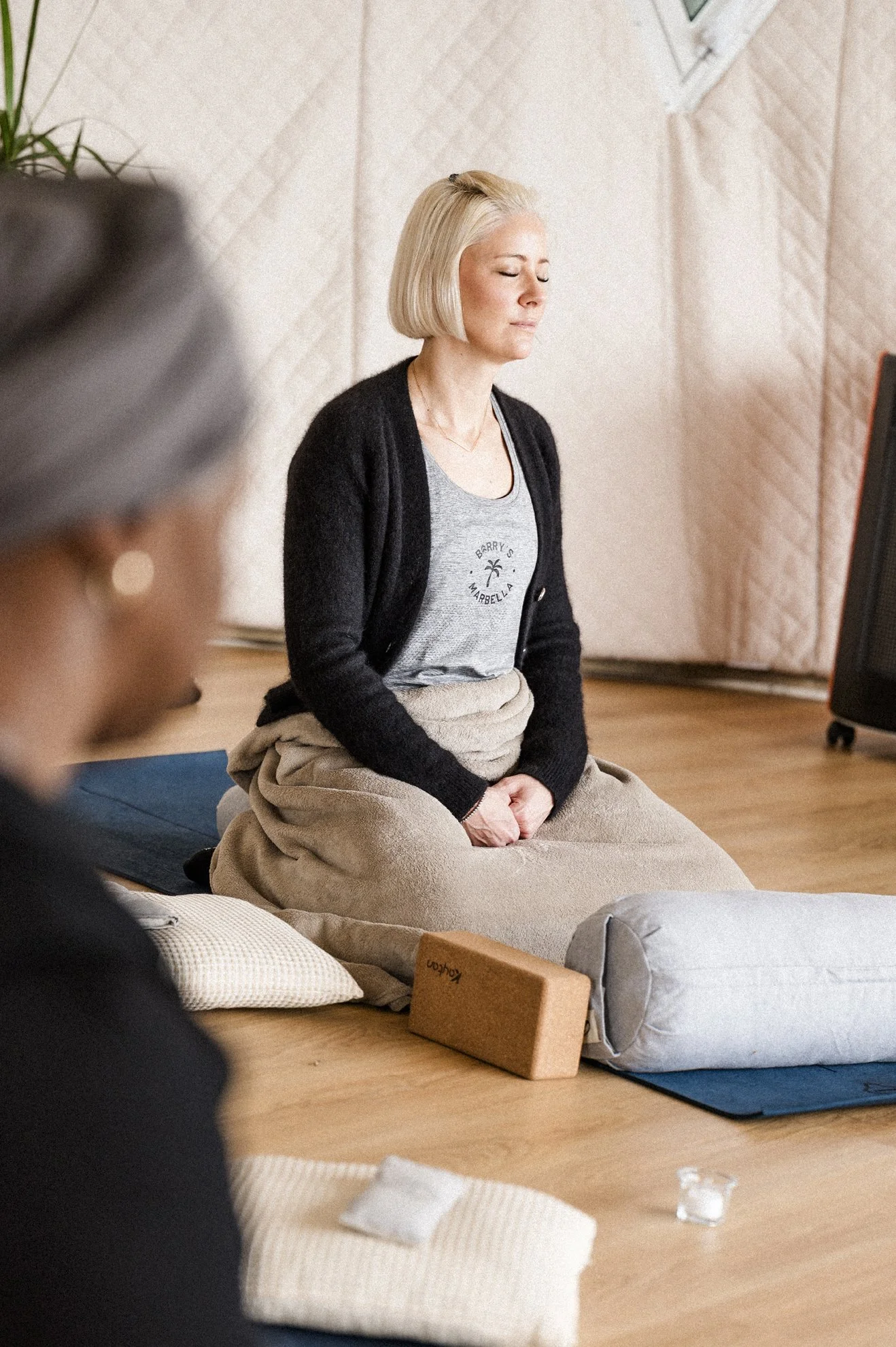 Eine blonde Frau sitzt mit geschlossenen Augen im Meditationsraum auf einer Matte, die Hände ruhen auf ihrem Schoß, umgeben von Kissen und Yogamatten.