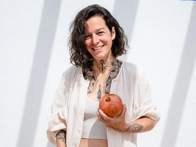 Eine lächelnde Frau mit Tattoos trägt ein weißes Oberteil und hält einen Granatapfel in der Hand vor einem weißen Hintergrund.