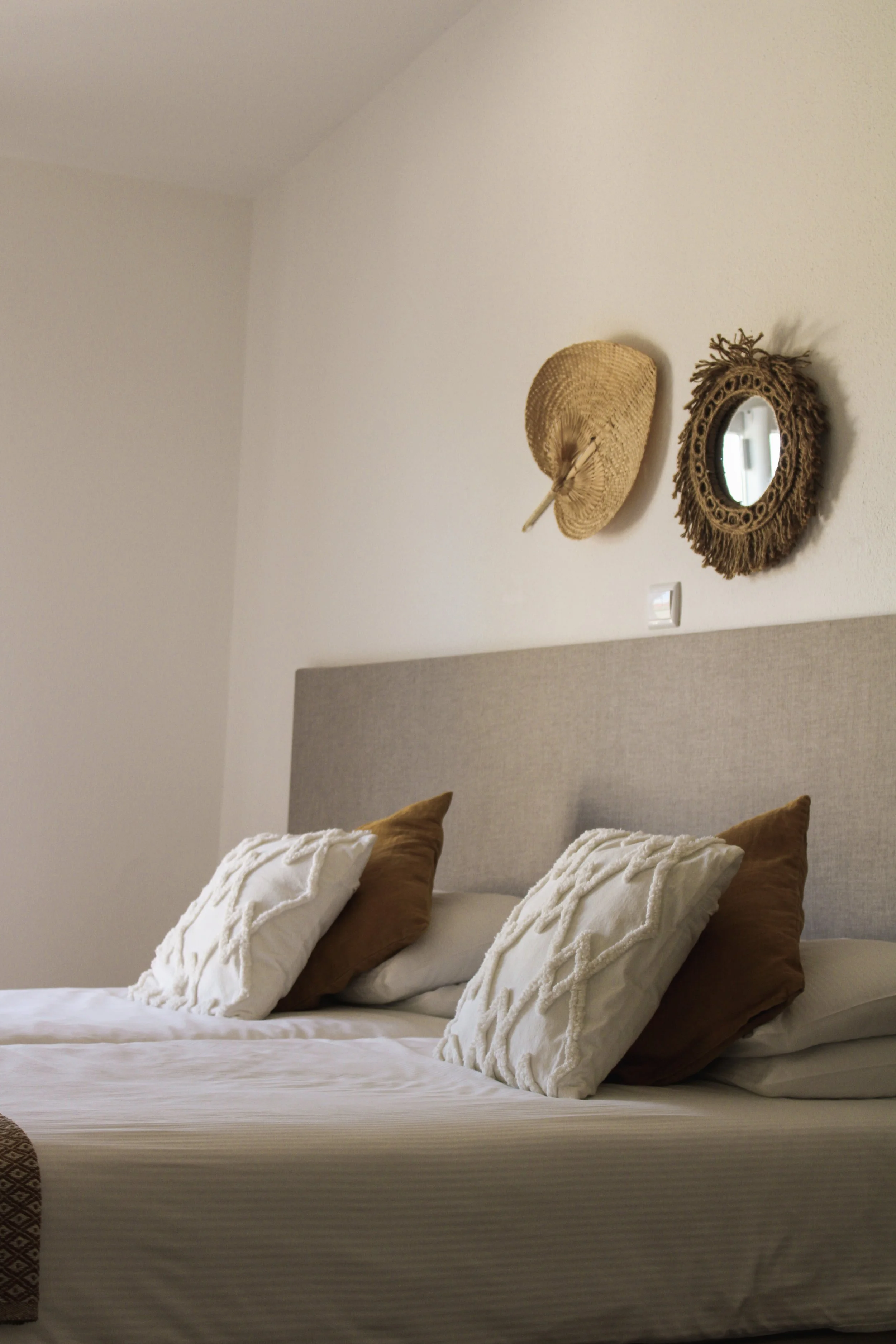 Bett mit weißen und braunen Kissen, an der Wand hängen zwei dekorative Spiegel, einer aus Korbmaterial und einer mit einem fransenbesetzten Rahmen, im Hintergrund eine Bettkopfseite
