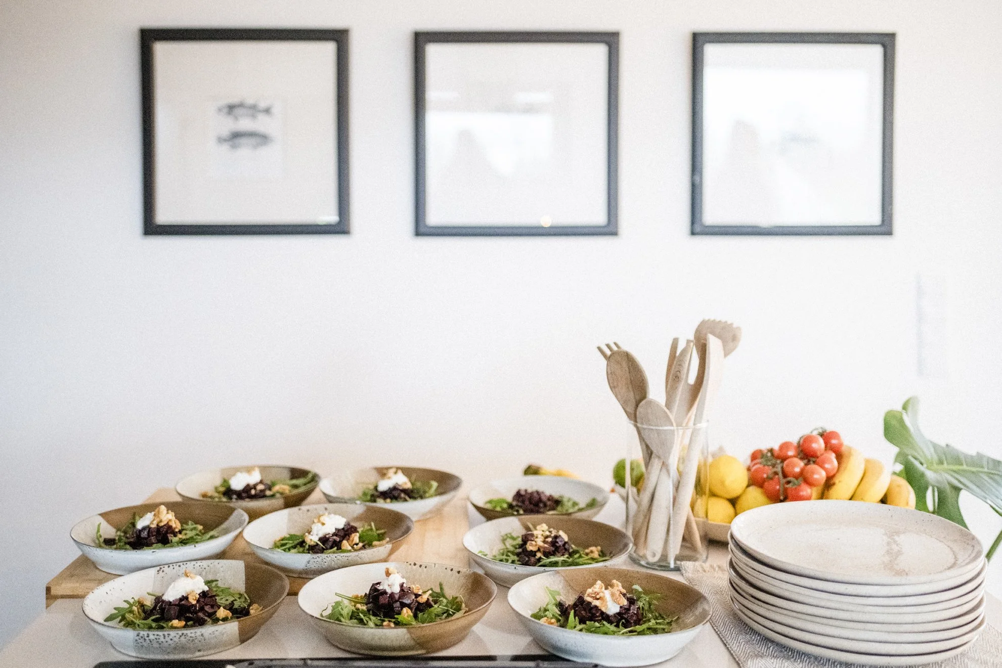 Verschiedene Schüsseln mit Salat und anderen Speisen auf einem Holztisch, im Hintergrund Obst und Küchenutensilien, an der Wand hängen drei Bilder