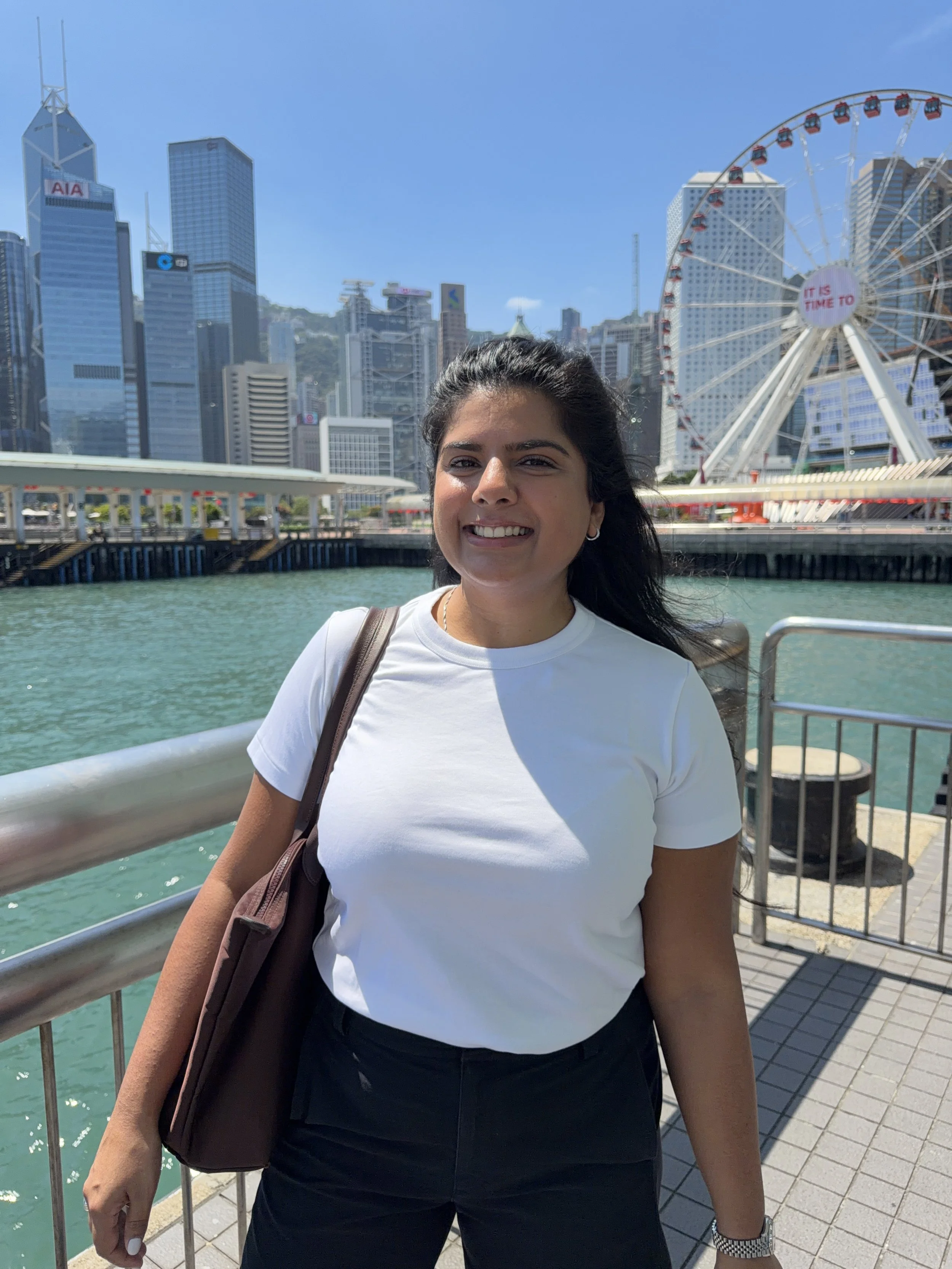 A young woman with dark hair, smiling, wearing a white t-shirt and black pants, standing by a railing near water with a city skyline and a Ferris wheel in the background.