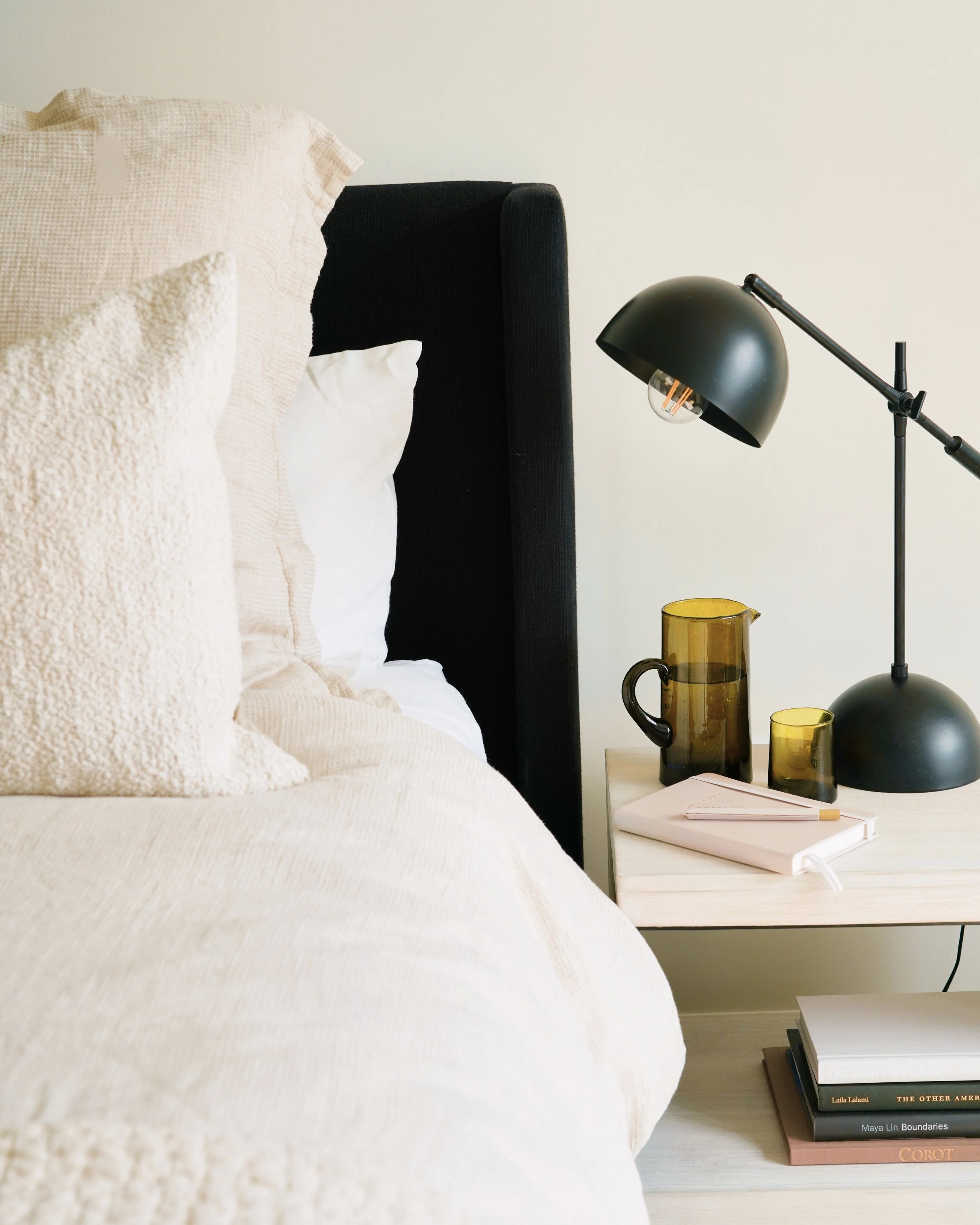 Close-up of a cozy bedroom corner showing a bed with textured cream-colored blankets and pillows, a black headboard, a white bedside table, a black adjustable desk lamp, and a stack of hardcover books in neutral colors.