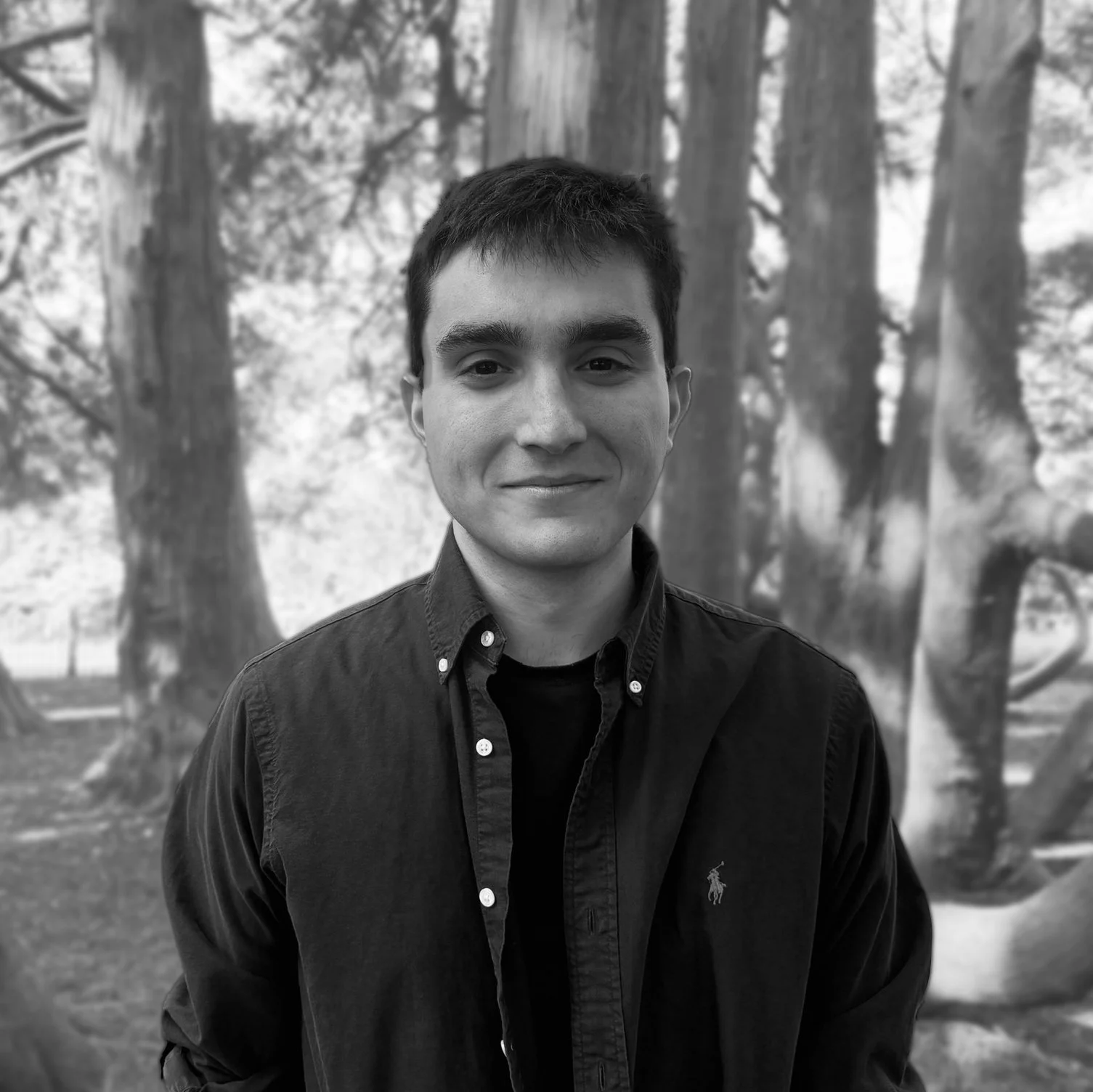 Michael Kats with short, dark hair, wearing a dark button-up shirt and smiling slightly, standing outdoors in front of trees with a blurred natural background.