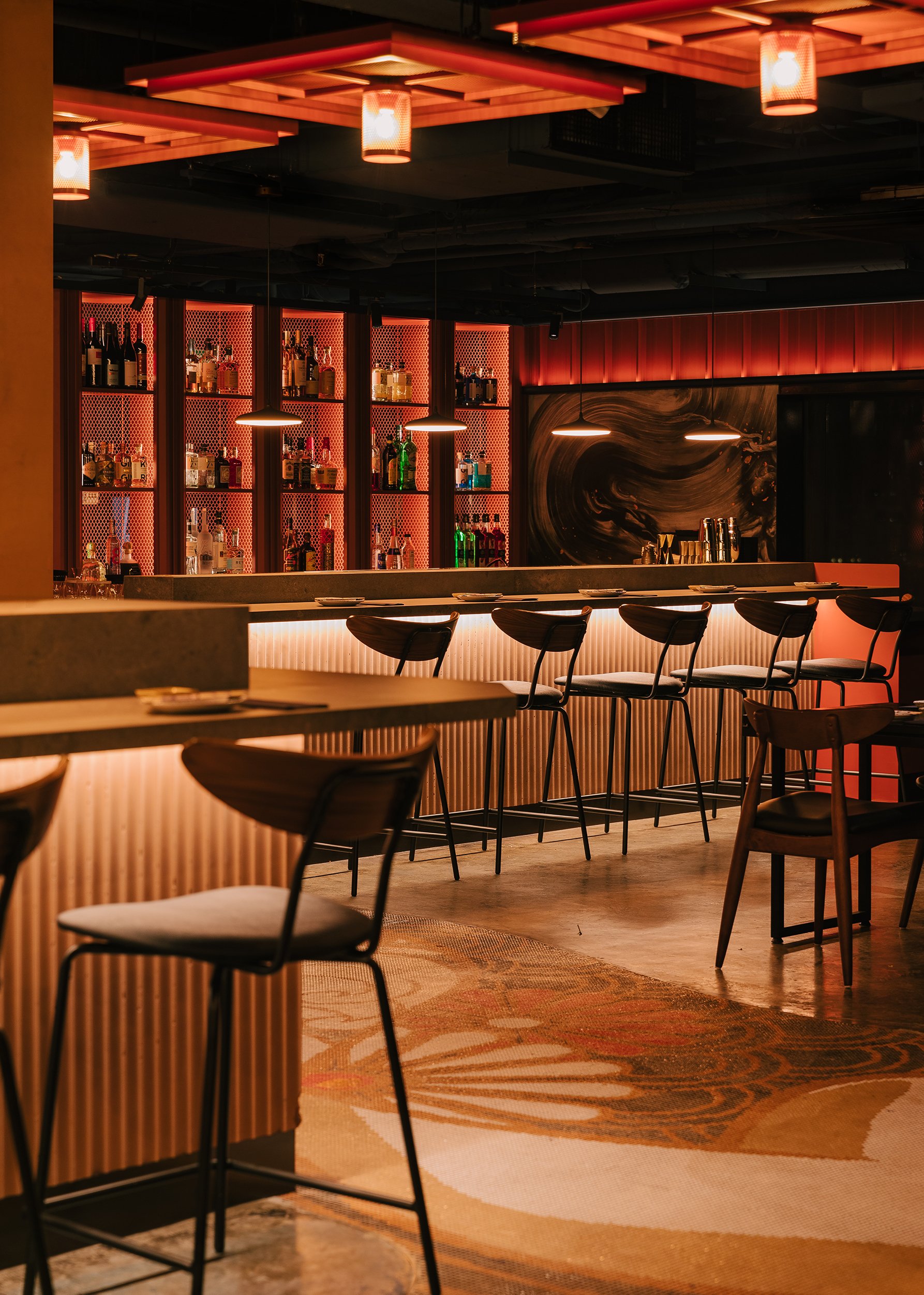 Modern bar with a wooden bar counter, black chairs, and liquor bottles on illuminated shelves. Warm lighting, artistic wall decor, and a patterned area rug create a cozy ambiance.