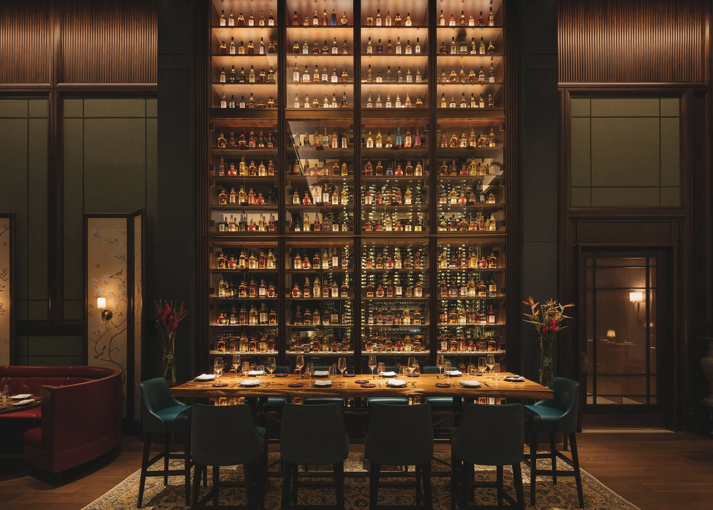 A luxurious restaurant interior with a tall, illuminated wooden shelf filled with bottles of alcohol, a long dining table set with wine glasses and plates, and chairs around the table. Dim lighting and elegant decor create a cozy atmosphere.