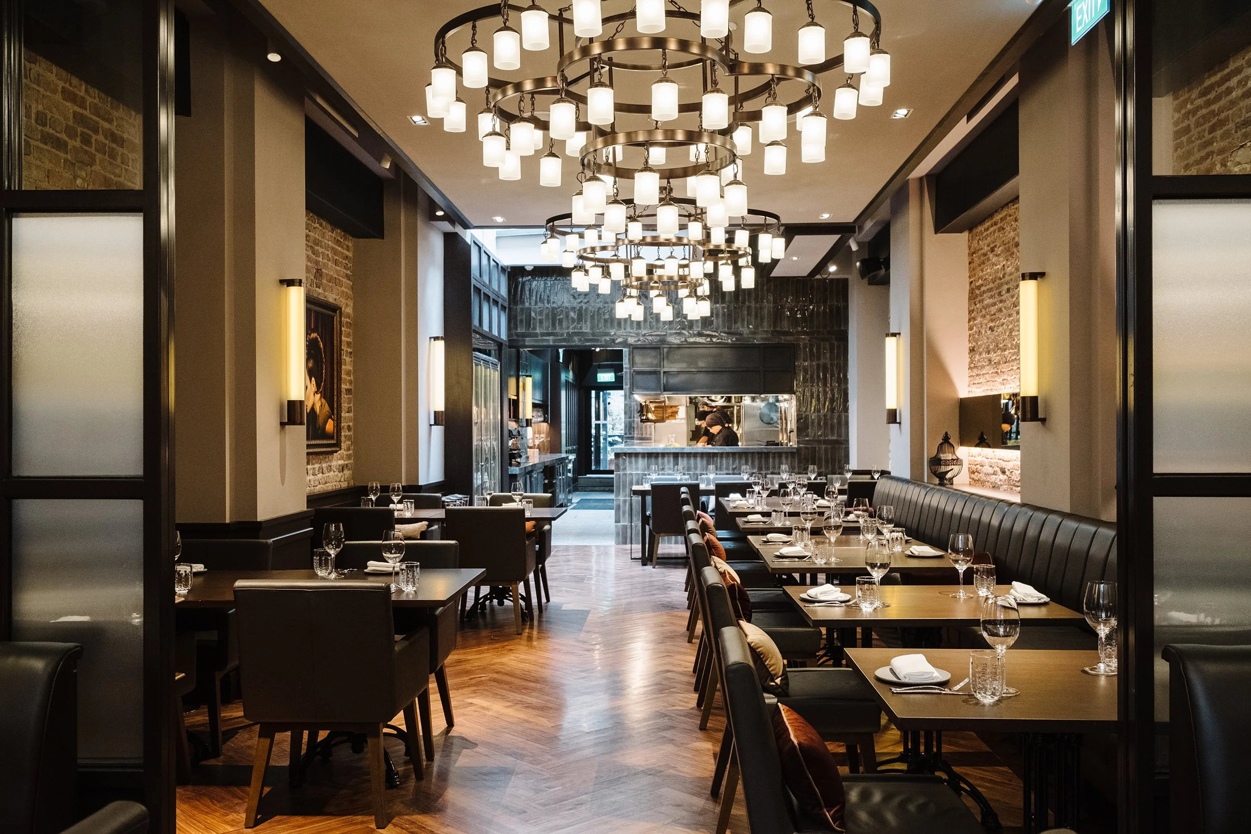 Interior of a modern restaurant with black chairs, wooden tables set with glasses and napkins, exposed brick walls, large chandelier with multiple lights, and a visible open kitchen area in the background.