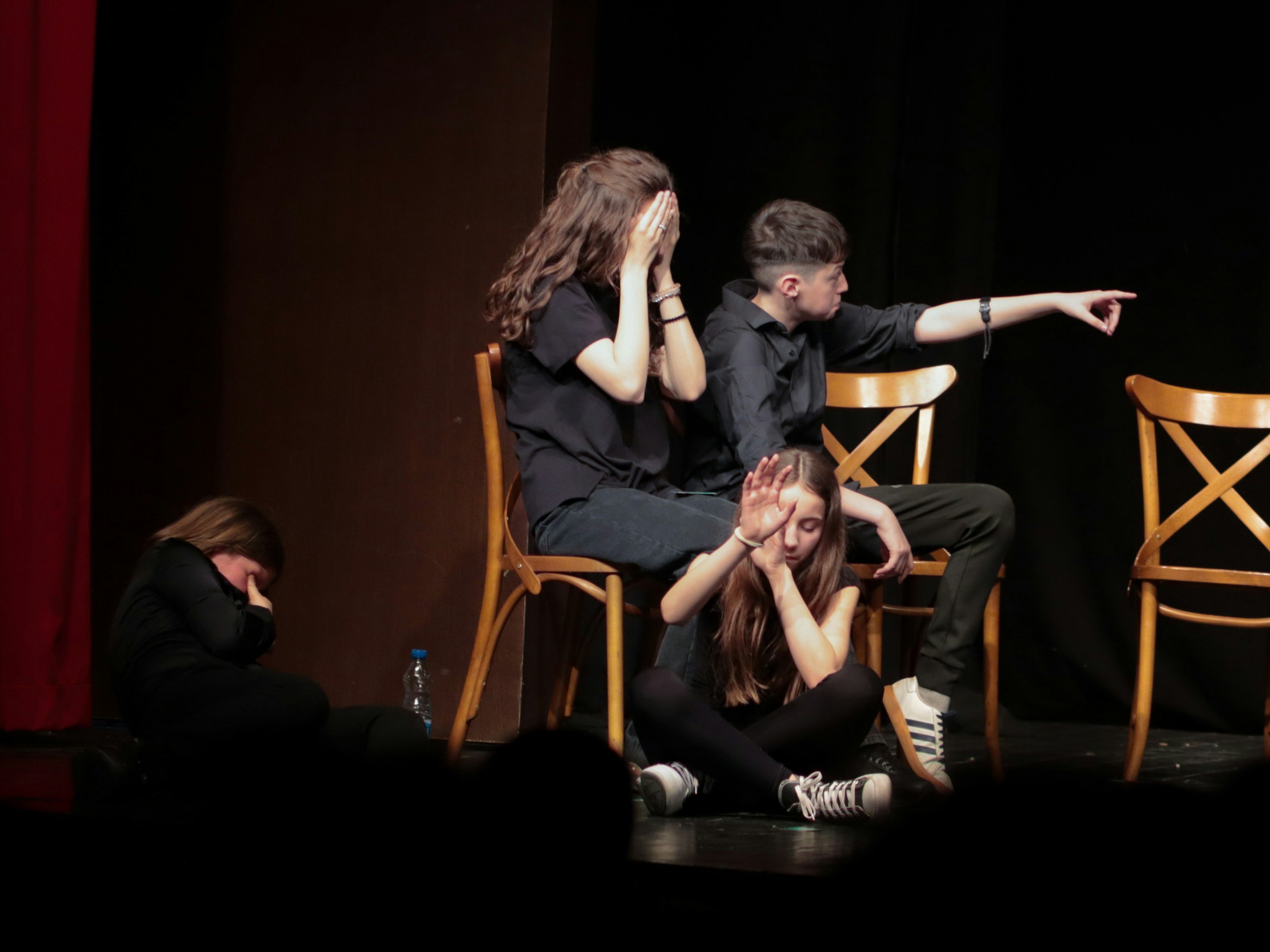 Gruppo di adolescenti seduti e sdraiati sul palco, alcuni con le mani sulla testa o sul viso, altri che puntano con le dita, in un ambiente scenico con sedie di legno e uno sfondo scuro.