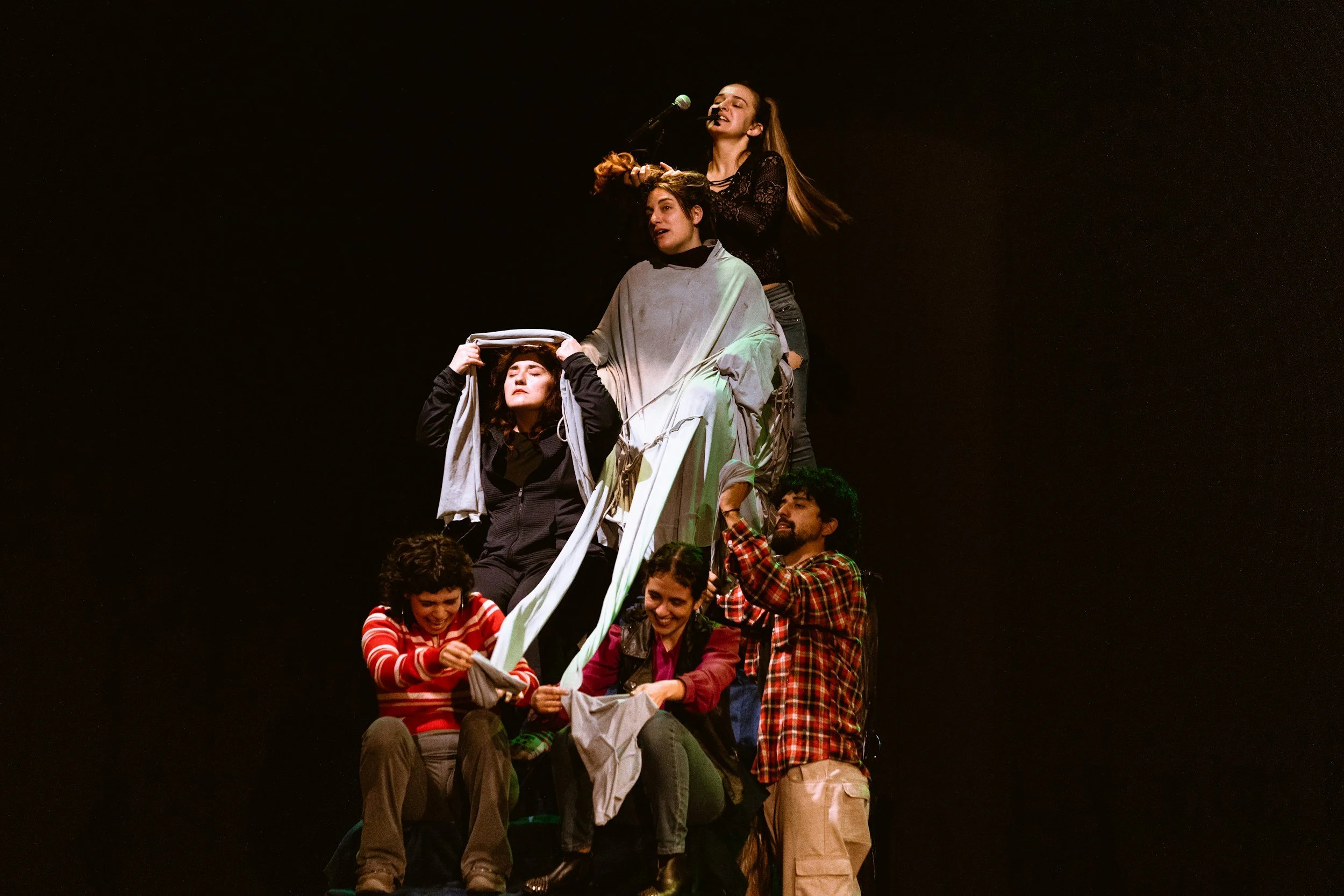 Gruppo di persone che formano una piramide umana durante una performance teatrale, con alcuni membri seduti e altri in piedi, sullo sfondo nero.