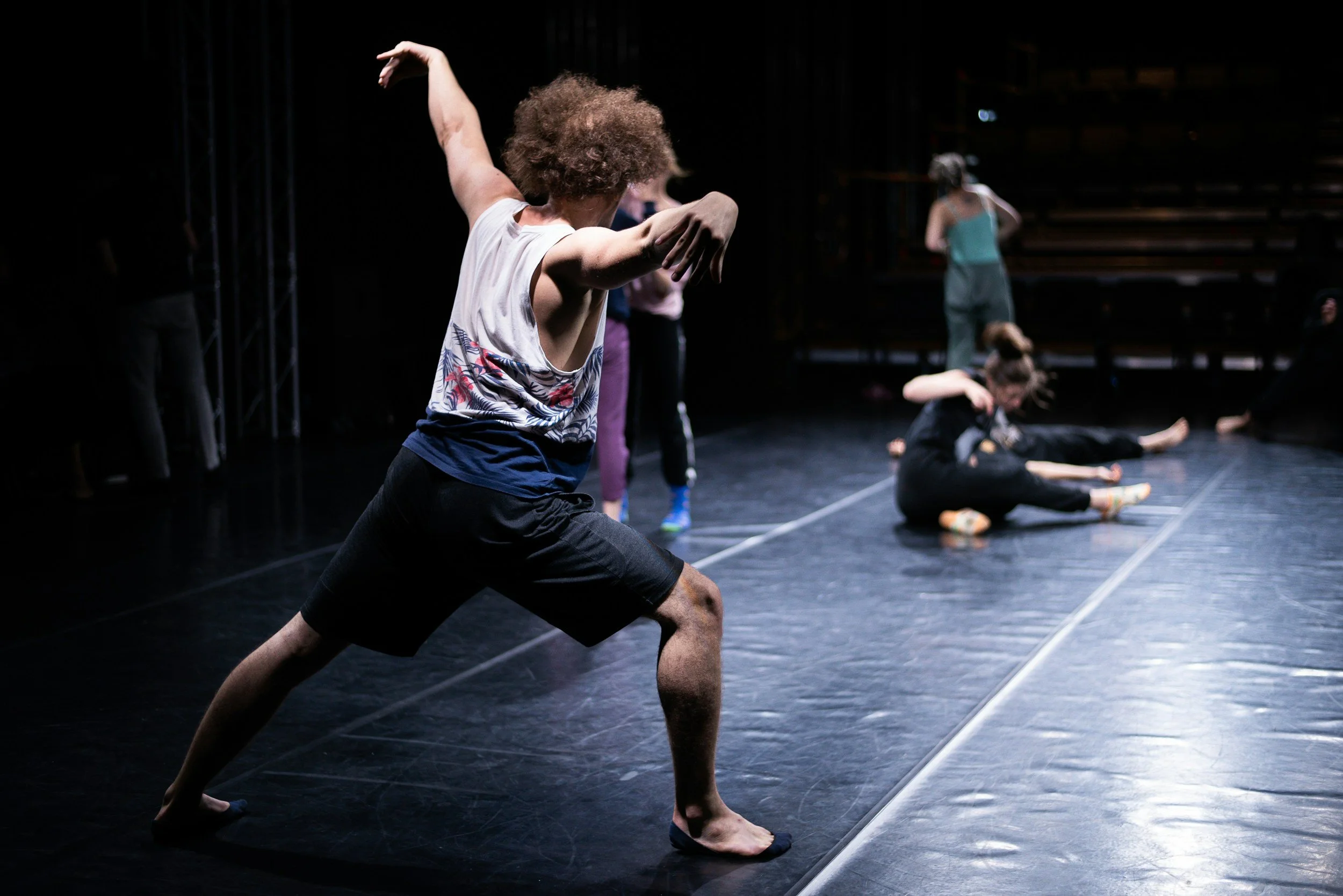Danza di ballerini in scena in un teatro buio, alcuni sono seduti sul pavimento e altri sono in posizione di stretching o movimenti di danza, con luci soffuse.