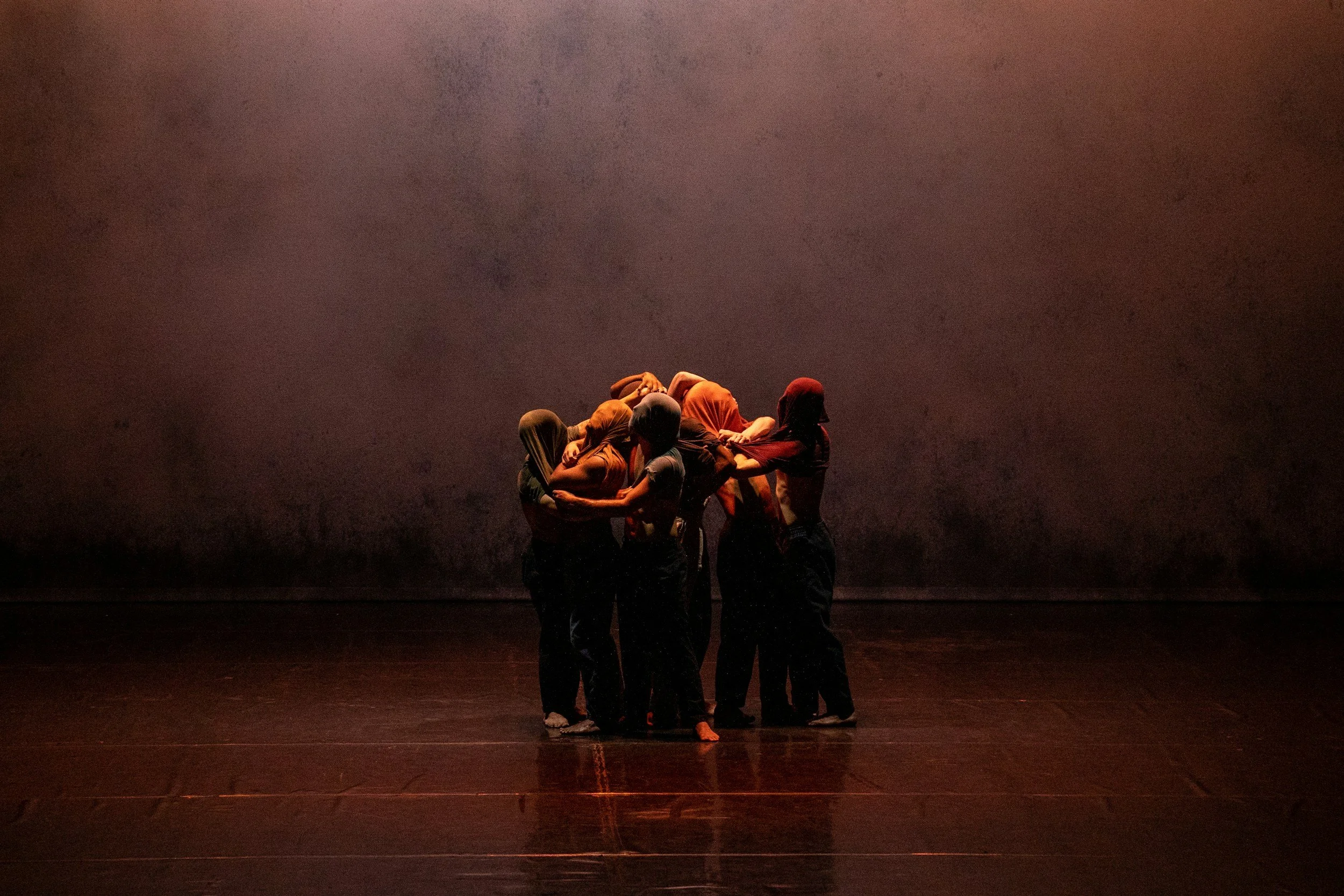 Un gruppo di persone con vestiti colorati si abbracciano e si stringono tra loro, formando una catena umana. Sono in piedi sul palco in un ambiente teatrale o di danza, con una parete scura come sfondo e luci soffuse.
