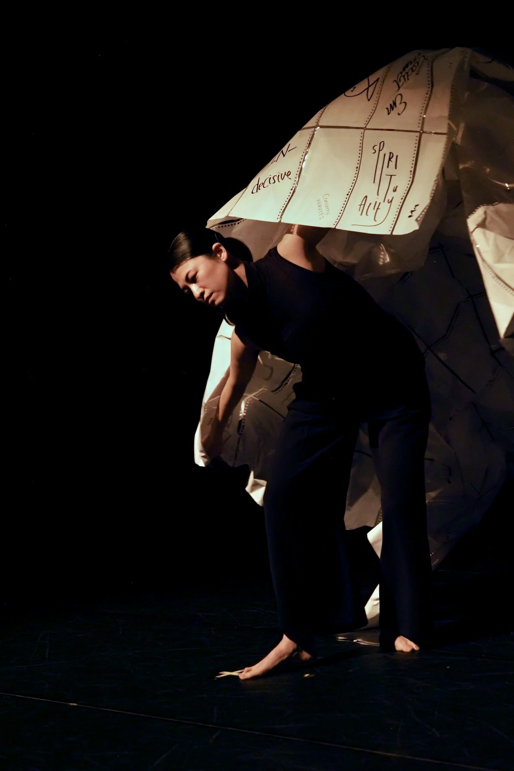 Una donna in abiti neri si piega in avanti su una scena di teatro, con un costume con scritte e disegni, in un'illuminazione scura.