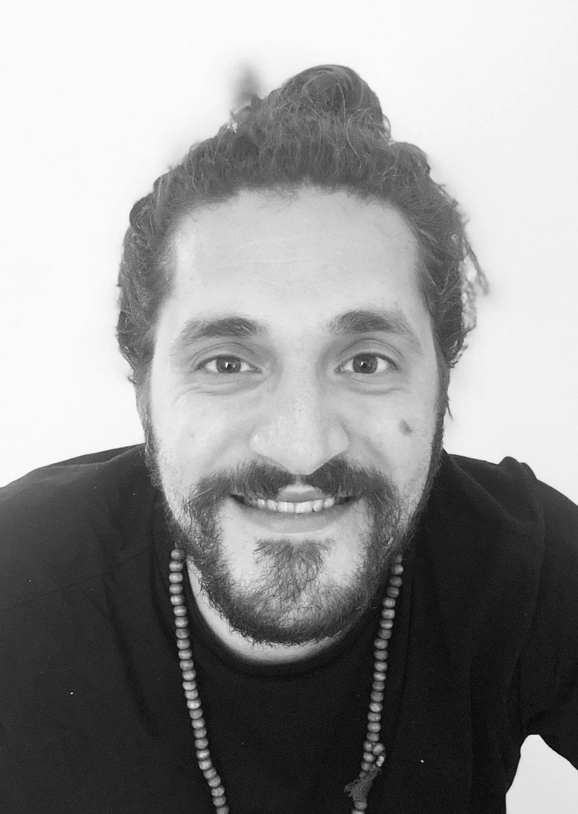 Ritratto di un uomo sorridente con barba e capelli raccolti, indossa una maglietta nera e una collana di perle. Sfondo bianco.