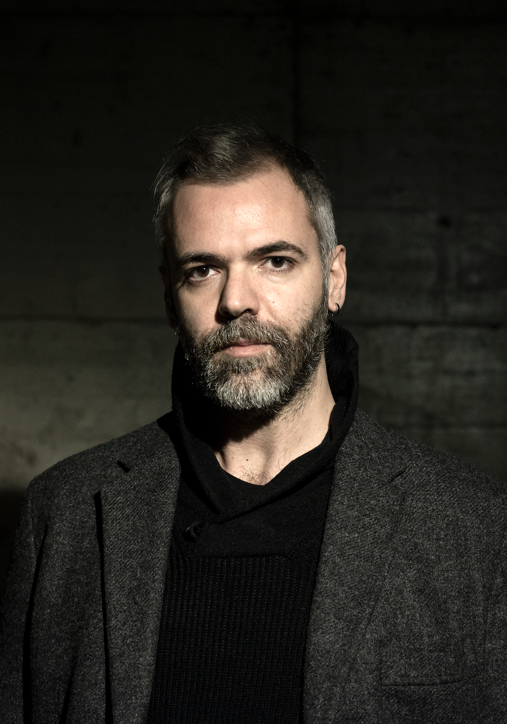 Ritratto di un uomo con barba e capelli castani, indossa un cappotto grigio e una felpa nera, sfondo scuro.