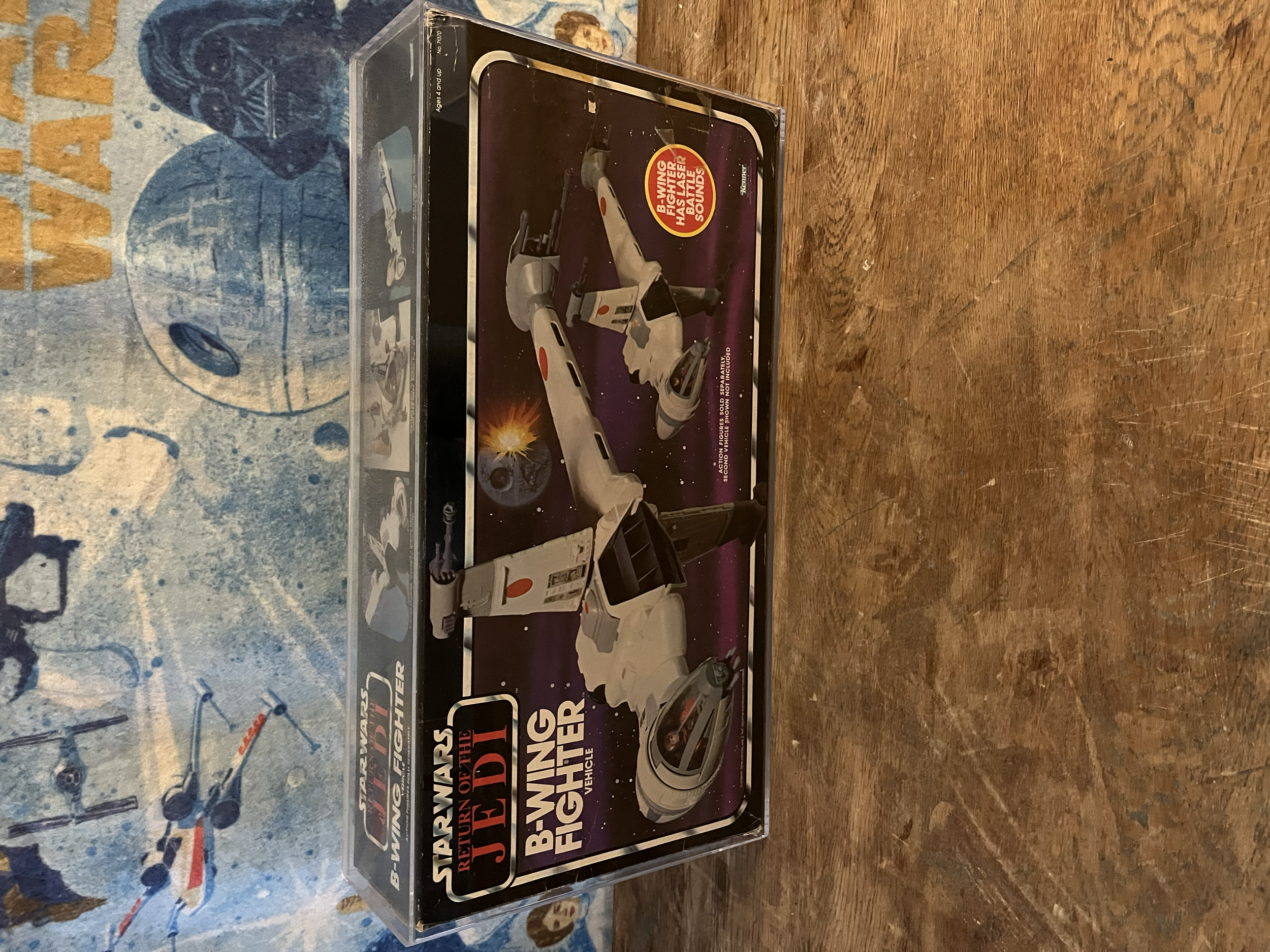 The image shows a vintage toy box with a space-themed design, featuring a model space shuttle and promotional text about the shuttle. It is placed on a wooden table, with a background featuring a blue and gray patterned surface.