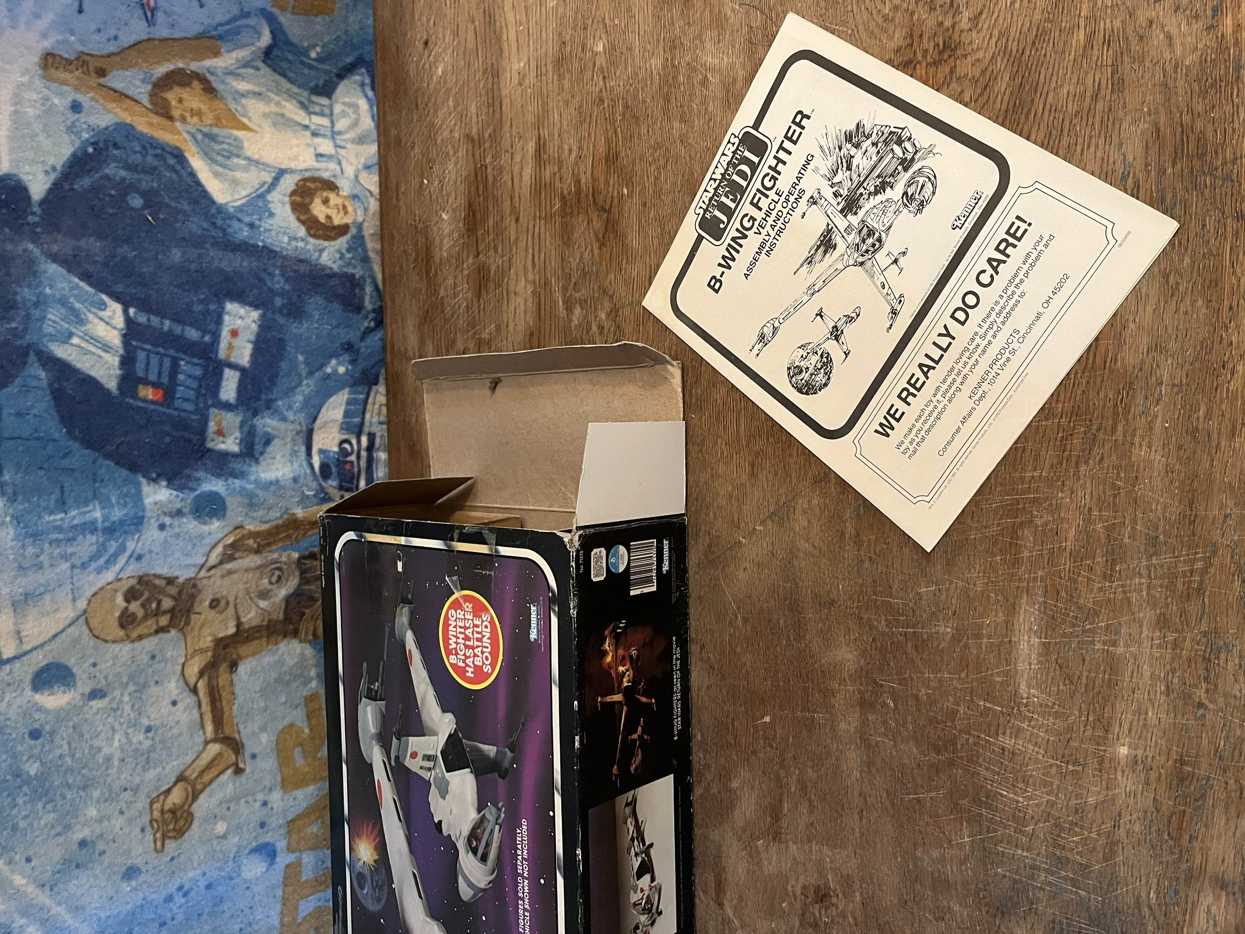 A vintage tabletop video game with packaging displaying a racing game, a B-wing fighter model, and game instructions beside a blue Star Wars-themed blanket with characters from the Star Wars series.