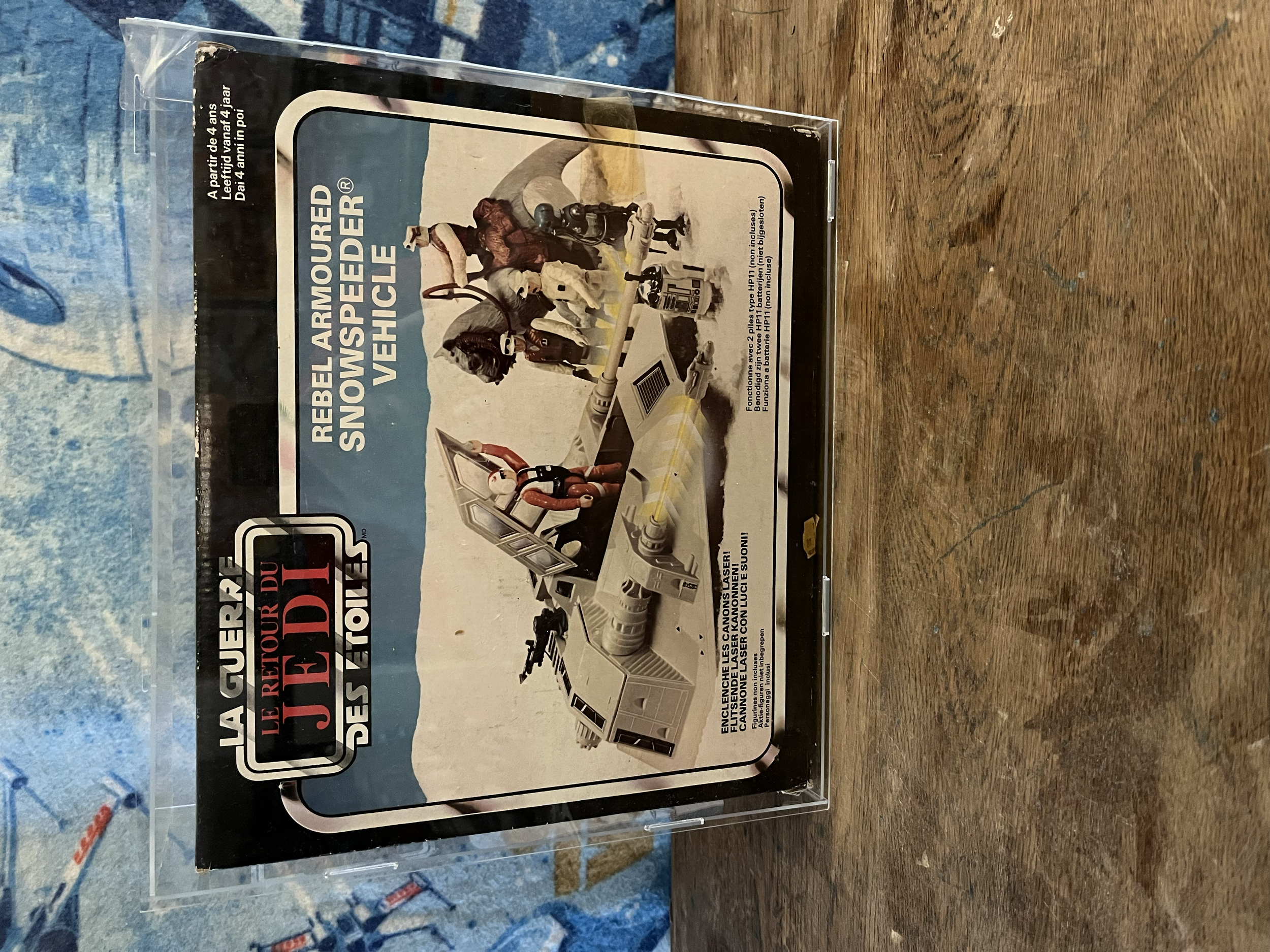 A sealed plastic package containing a toy or model of a robot or machine with a crew member figure on top, positioned on a wooden surface with a blue patterned cloth in the background.
