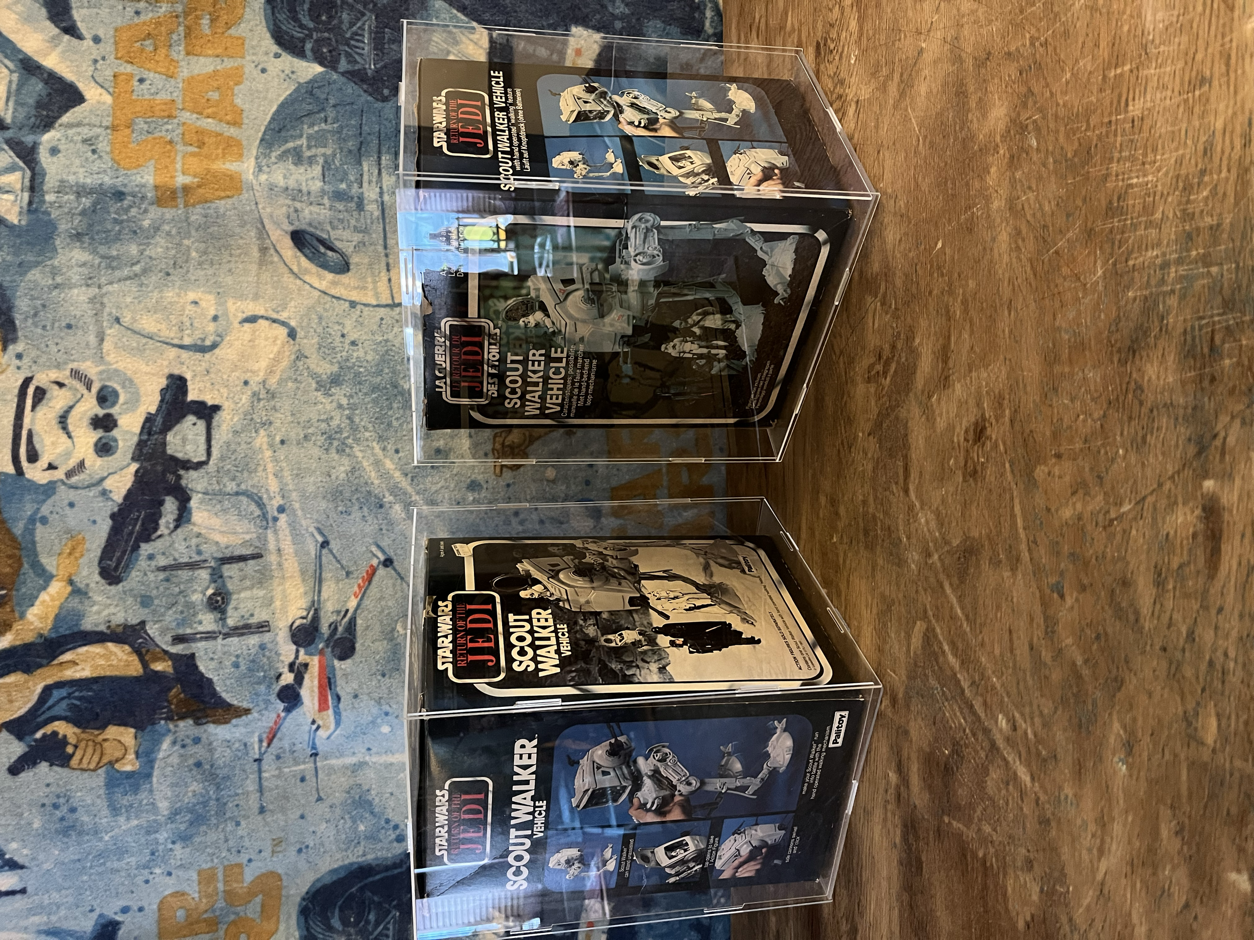 A clear display case containing an action figure of a Scout Walker from Star Wars, with a box packaging behind it showing the figure and text related to Star Wars: Return of the Jedi.