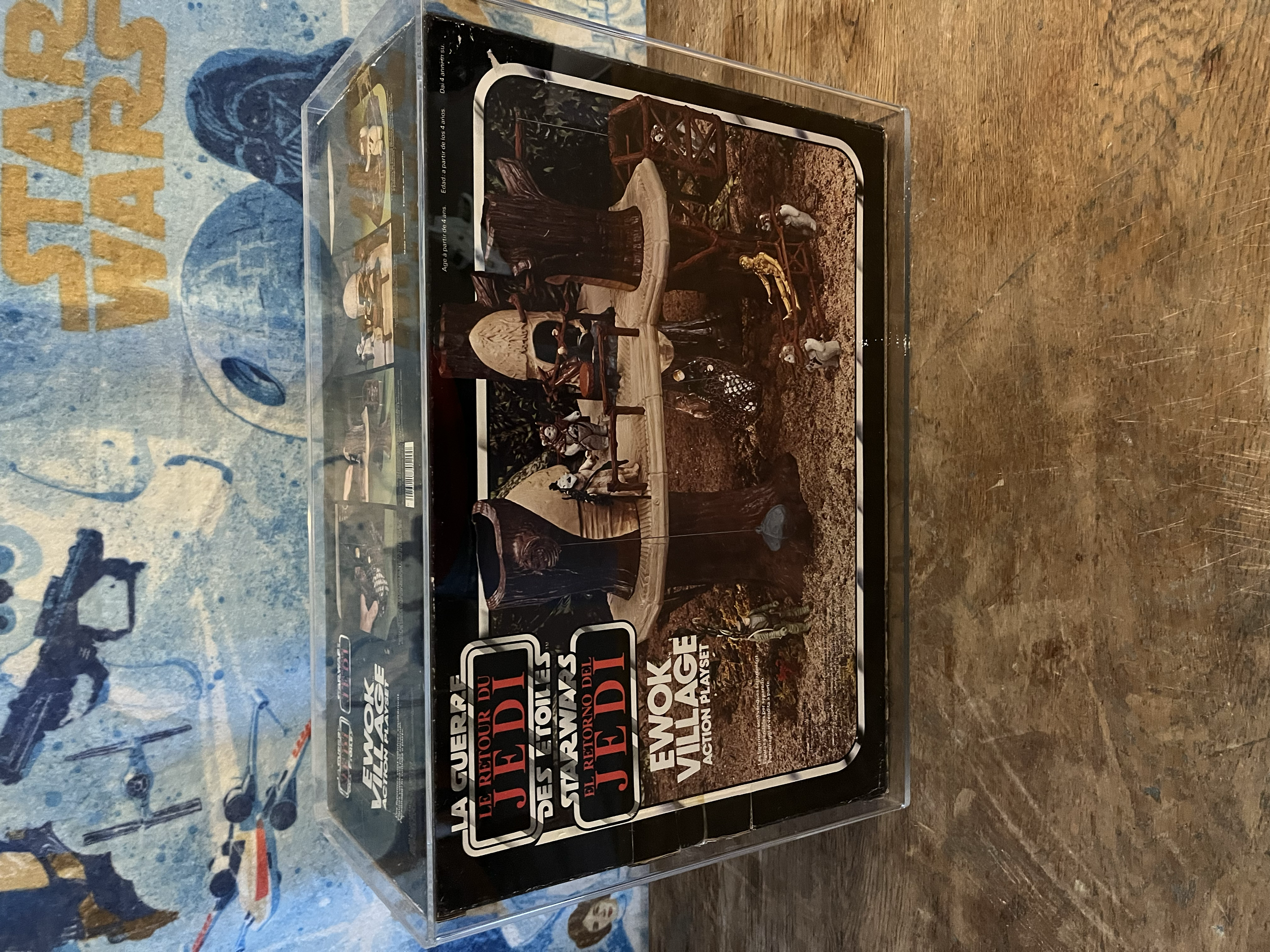 A clear plastic display case containing several black and orange labels with red and white text. The labels feature text in French and appear to be related to a product or promotional material. The case sits on a wooden surface with a painted surface
