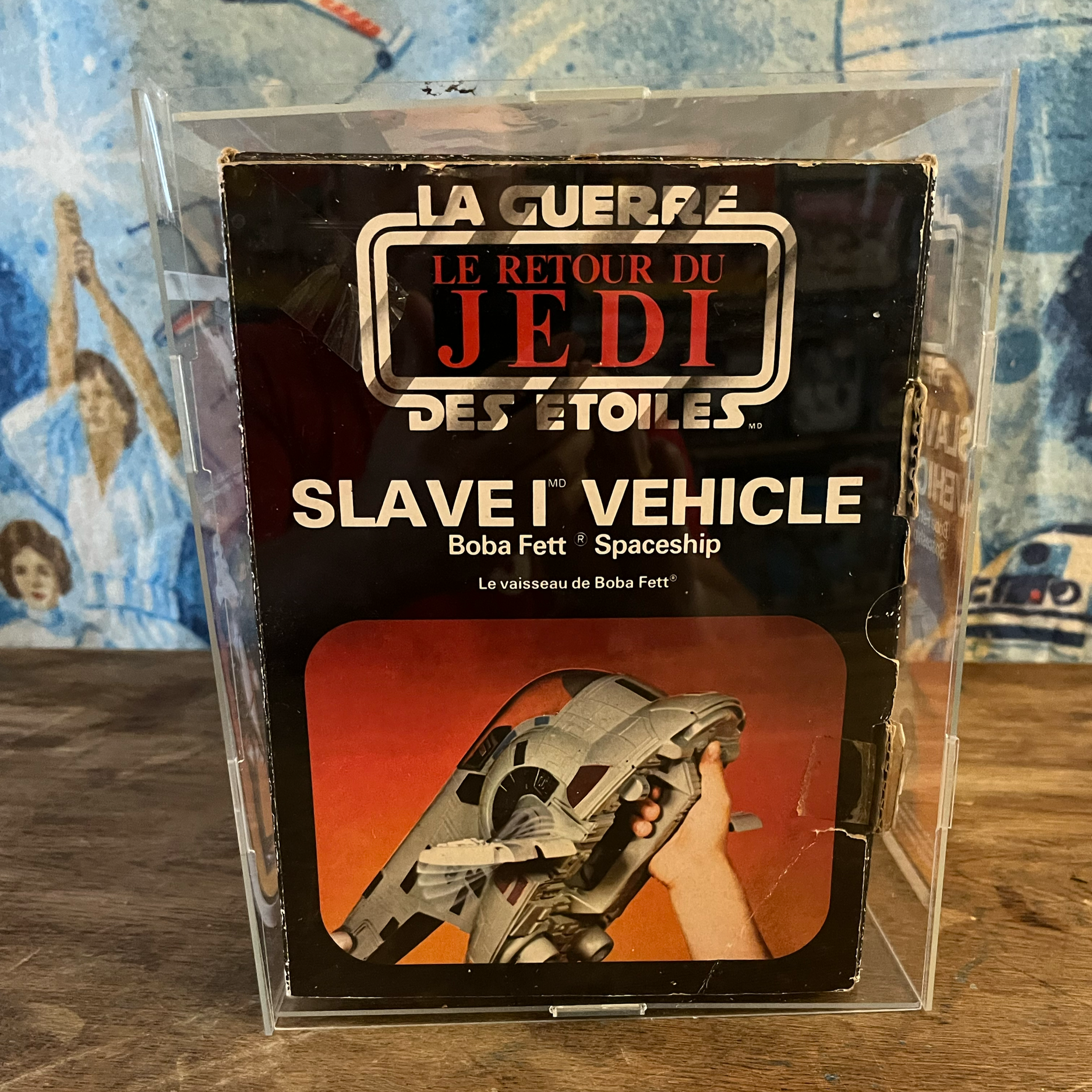 Close-up of a vintage Star Wars toy box for 'Le Retour du Jedi' featuring a slave I vehicle from Boba Fett, with a person holding it. The background includes a wooden surface and a blue patterned cloth with characters.