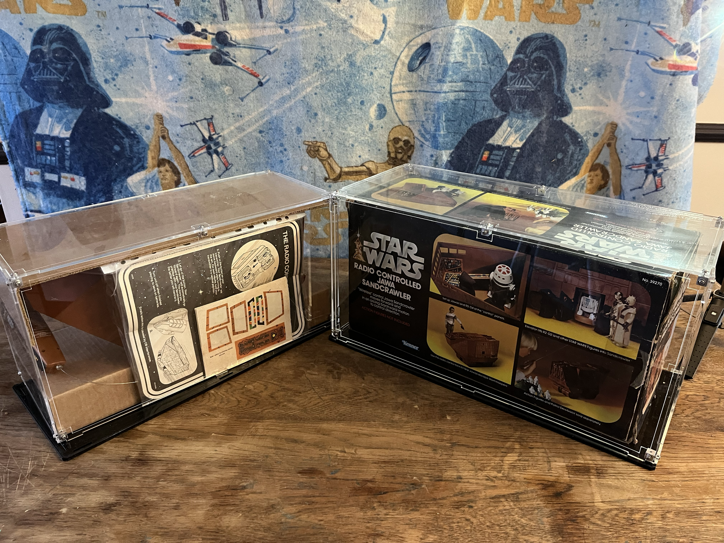Star Wars themed collectibles in clear display cases on a wooden table, with a vintage Star Wars comic in one case, a Kenner Star Wars radio-controlled Jawa Sandcrawler in another, and a Star Wars poster in the background.