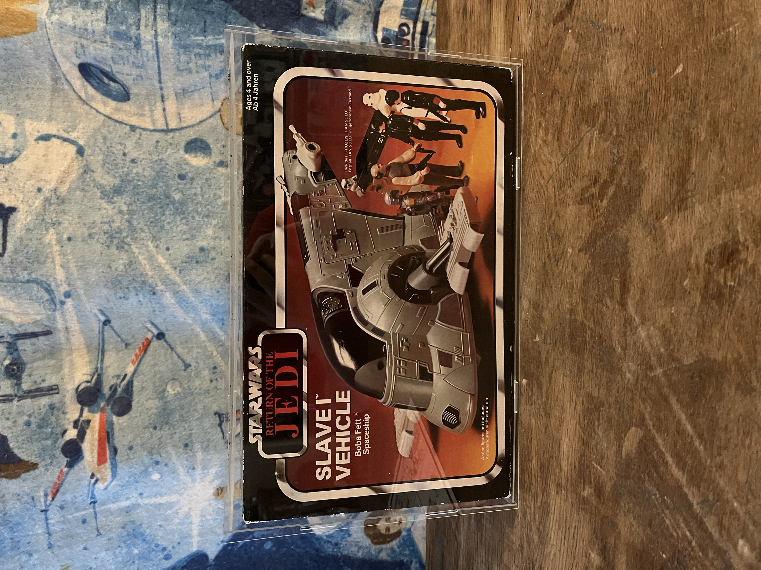 The image shows a box of the Star Wars: Return of the Jedi toy vehicle with an illustration of the vehicle and characters from the movie on it, placed on a wooden surface with a blue patterned background.