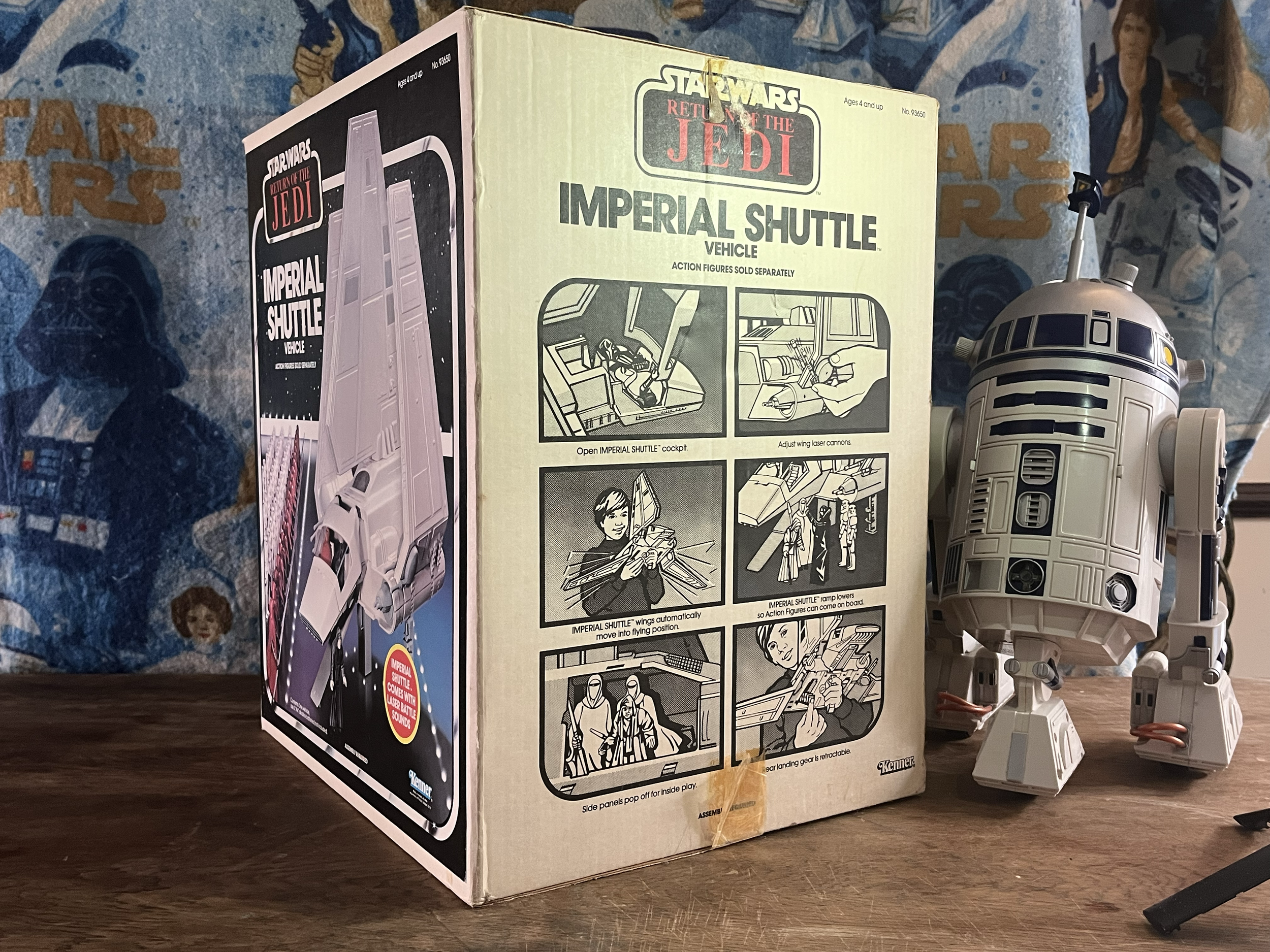 A boxed model of an Imperial Shuttle from Star Wars, with assembly instructions and images on the side. The box is placed on a wooden surface with a Star Wars themed blanket in the background. To the right of the box, there is a R2-D2 toy.