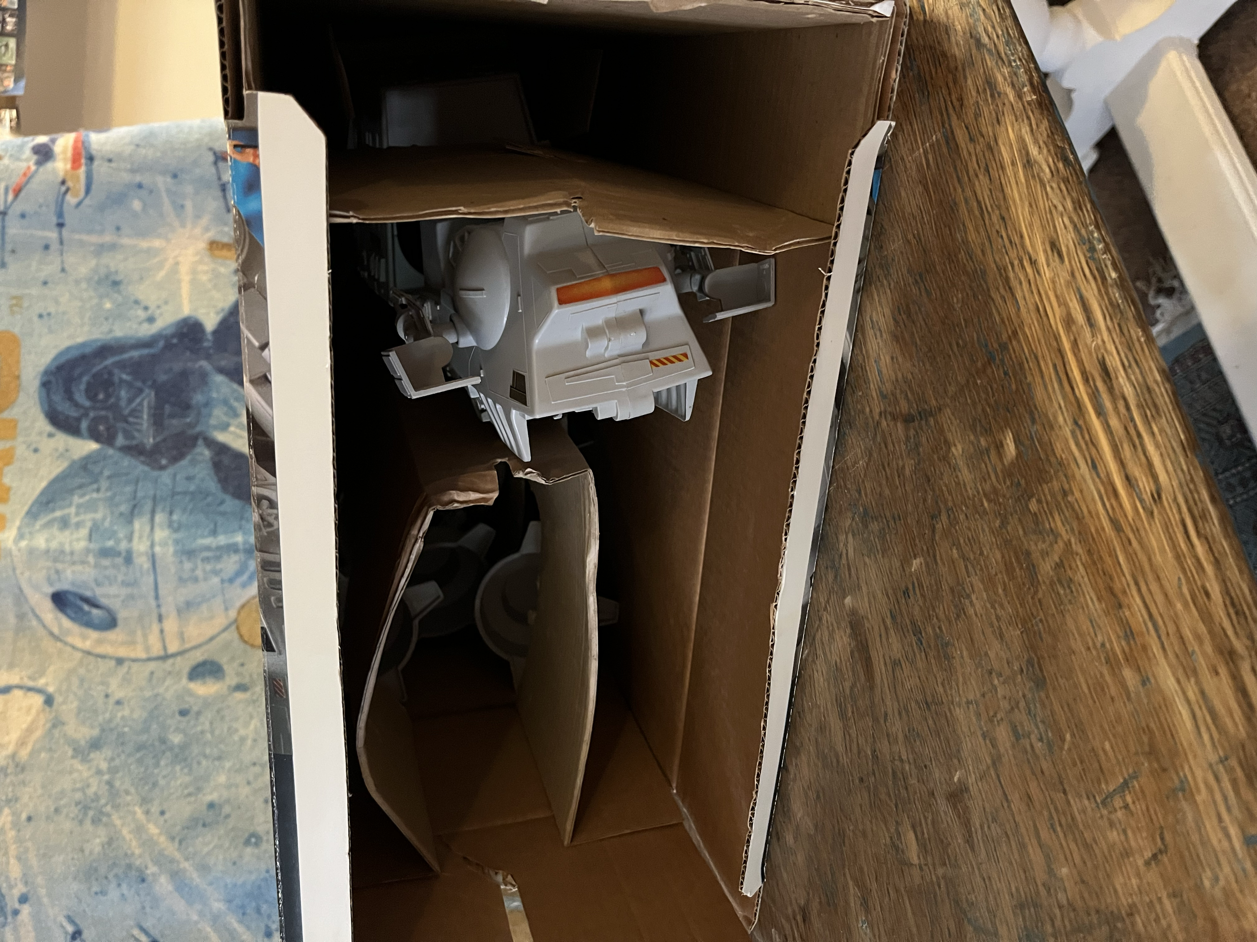 A cardboard shipping box with an empty wooden shelf inside, placed on a wooden surface, with a partially visible map or poster on the left side.