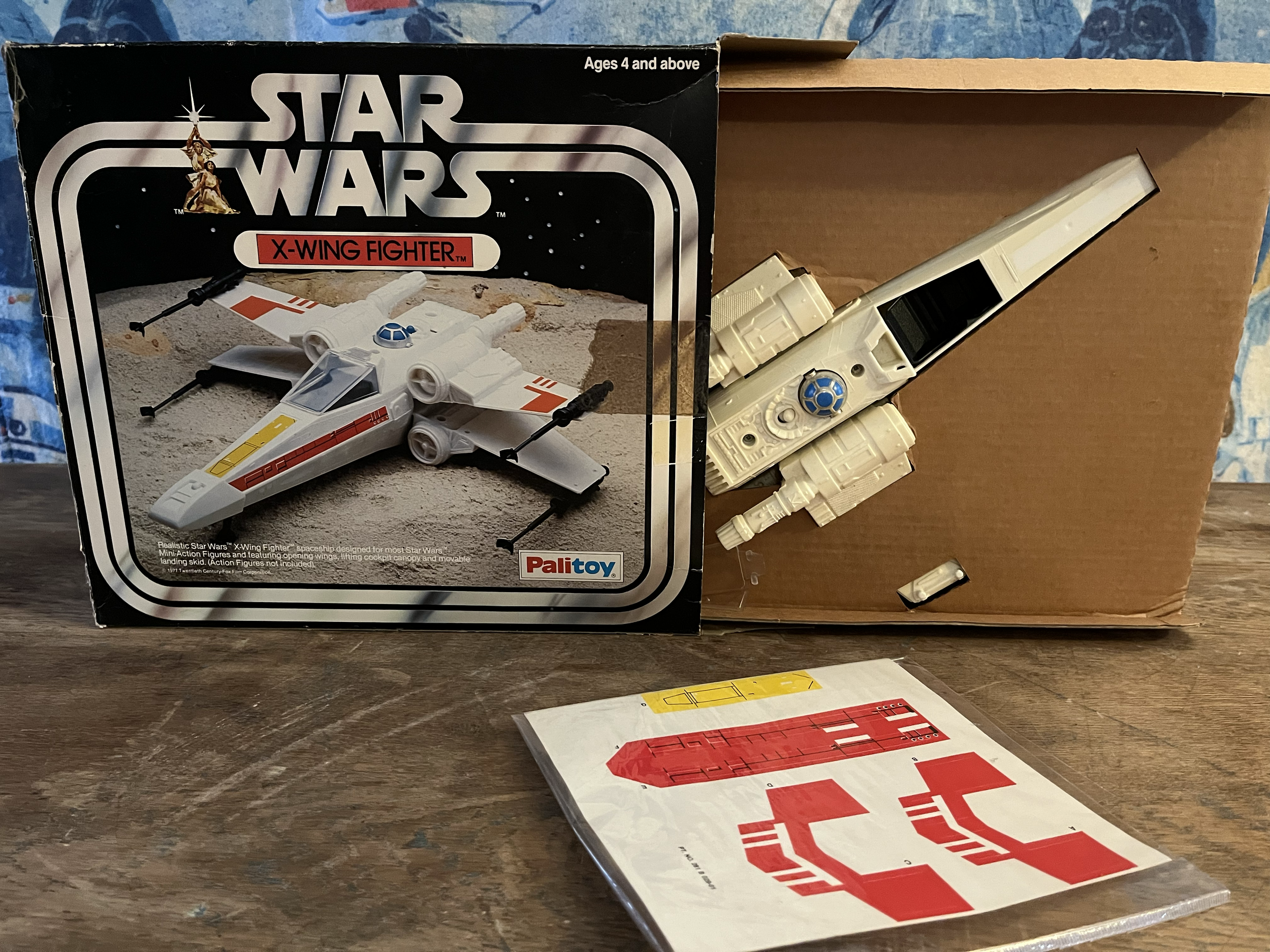 A vintage Star Wars X-Wing Fighter model kit box with an illustration of the spaceship on the front, showing a white spacecraft with red and blue markings, along with some unassembled model parts and stickers on a wooden table.