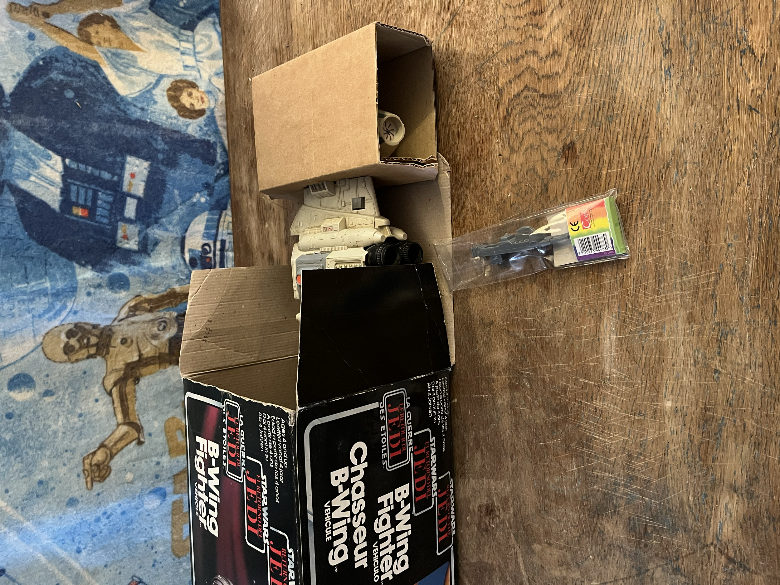 A tabletop with a Star Wars spaceship model inside a cardboard box, a black box labeled 'Star Wars Bubble Shield Fighters,' and a small plastic package with a green, red, and yellow label.