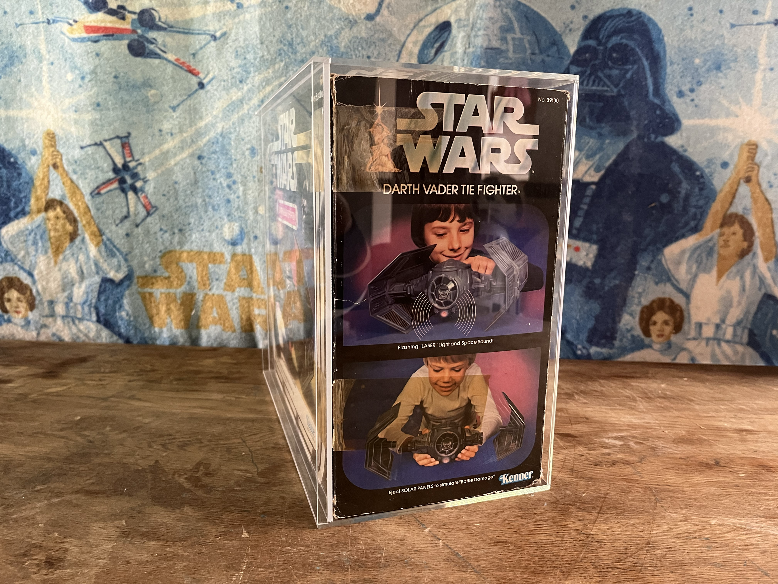 A vintage Star Wars Darth Vader Tie Fighter toy with solar panels and space sound features, displayed on a wooden surface against a Star Wars-themed background.