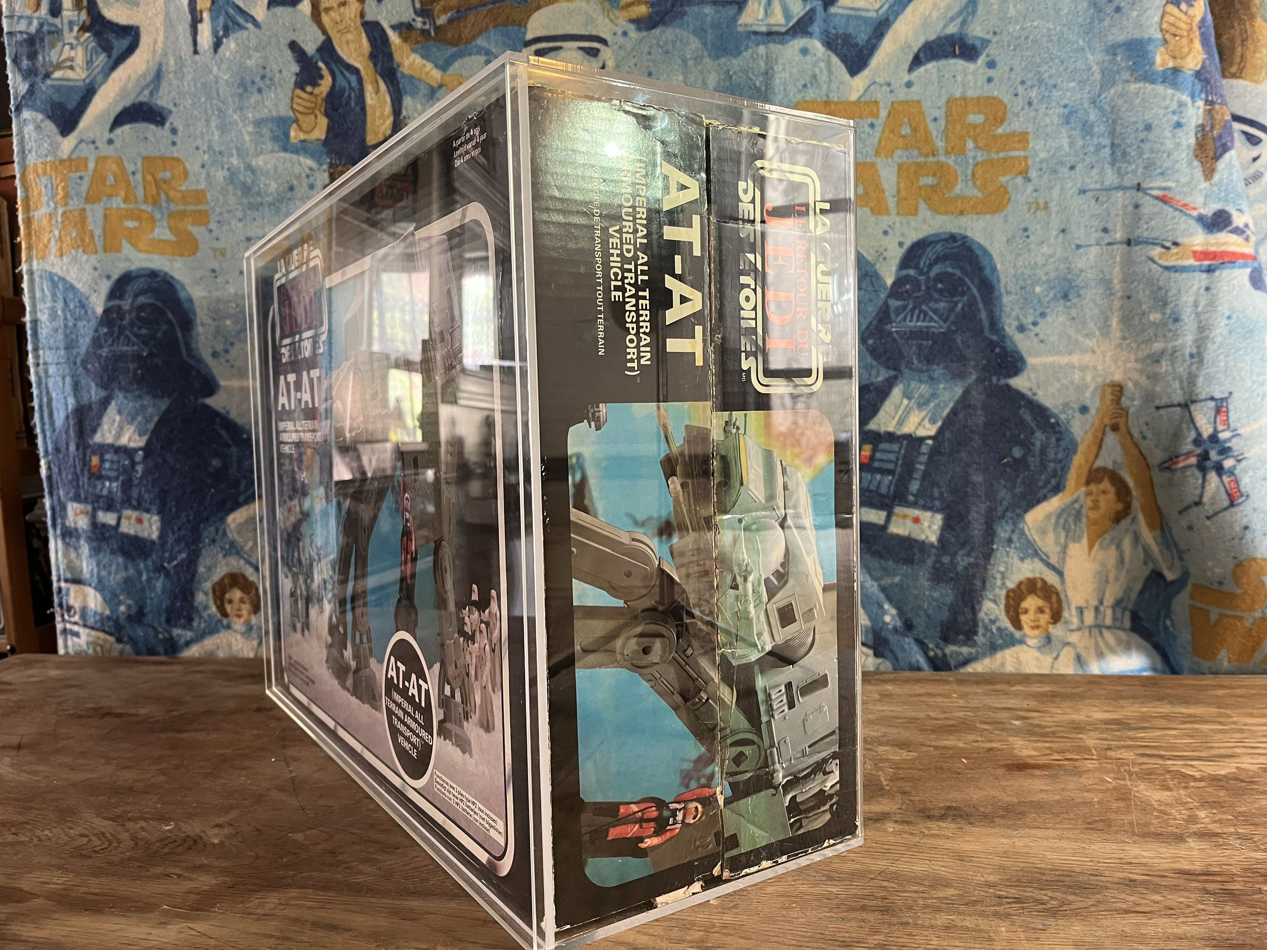 A clear plastic display case on a wooden table, showcasing a vintage Star Wars toy vehicle from the Imperial All Terrain Transport Vehicle line, with a Star Wars-themed cloth featuring characters like Darth Vader in the background.