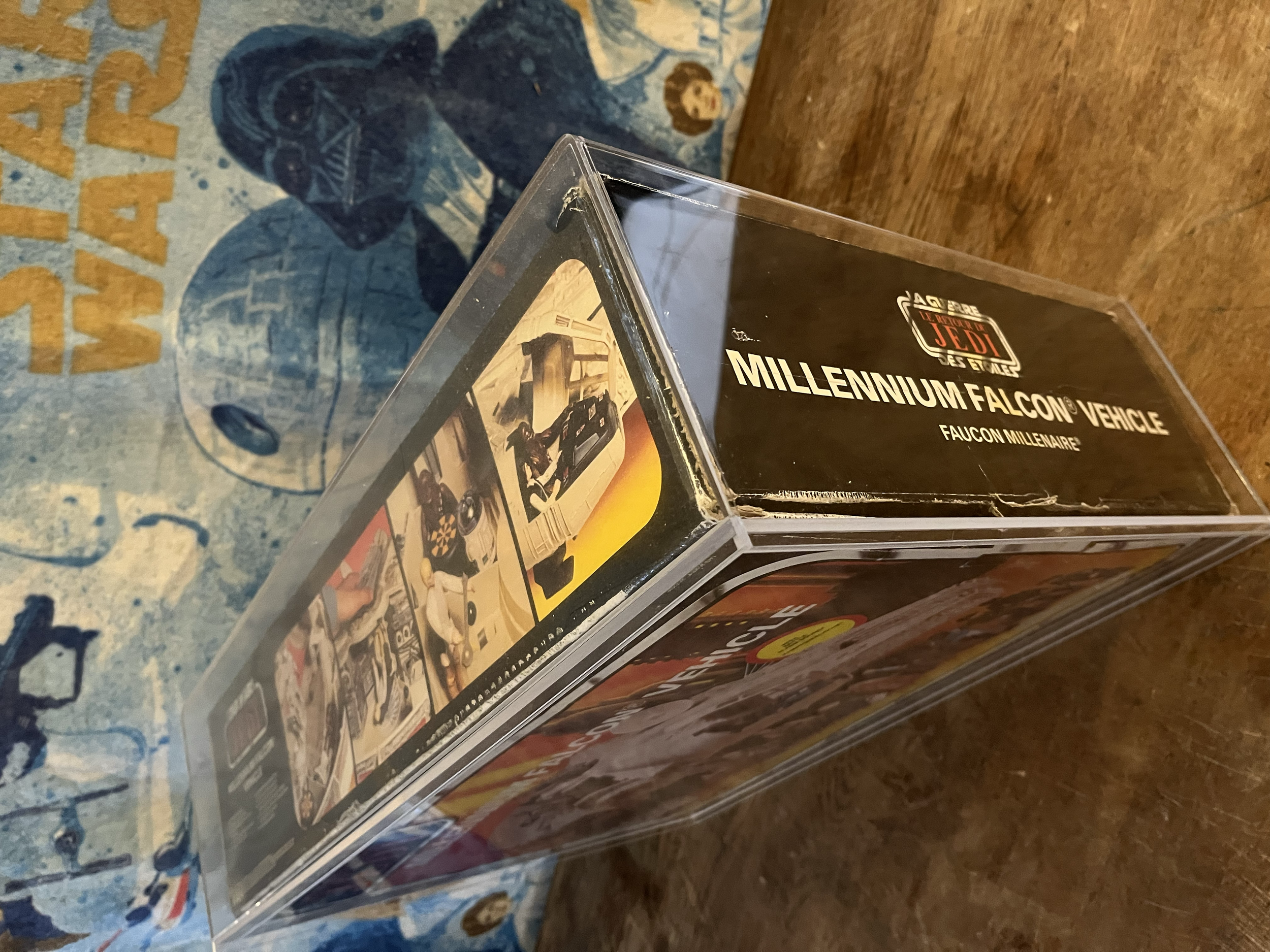 A vintage Star Wars Millennium Falcon vehicle in a plastic case with artwork and logos, resting on a wooden surface with a Star Wars-themed mat nearby.