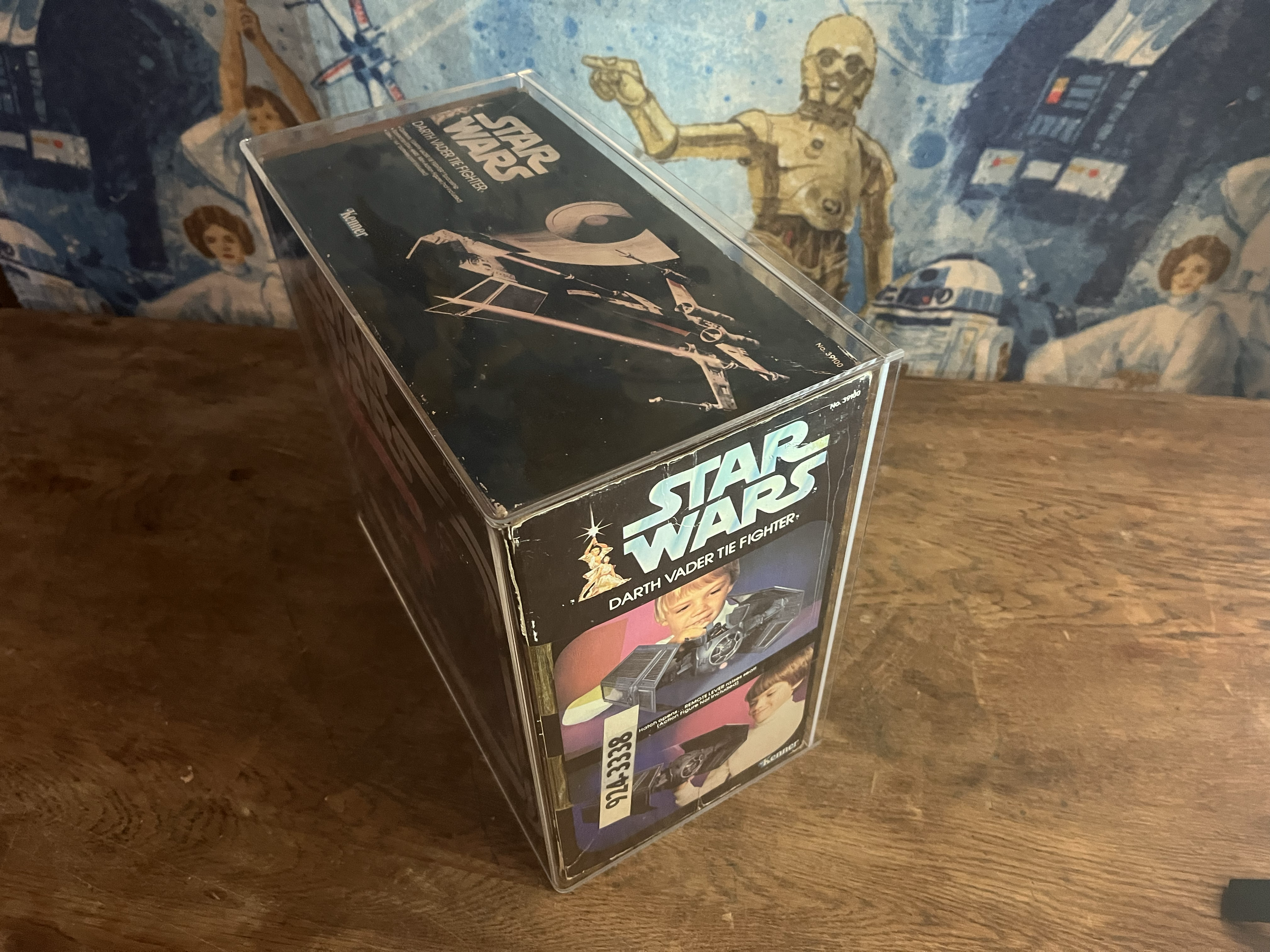 Close-up of a vintage Star Wars action figure box featuring Darth Vader and other characters, placed on a wooden surface with a star wars bedspread in the background.