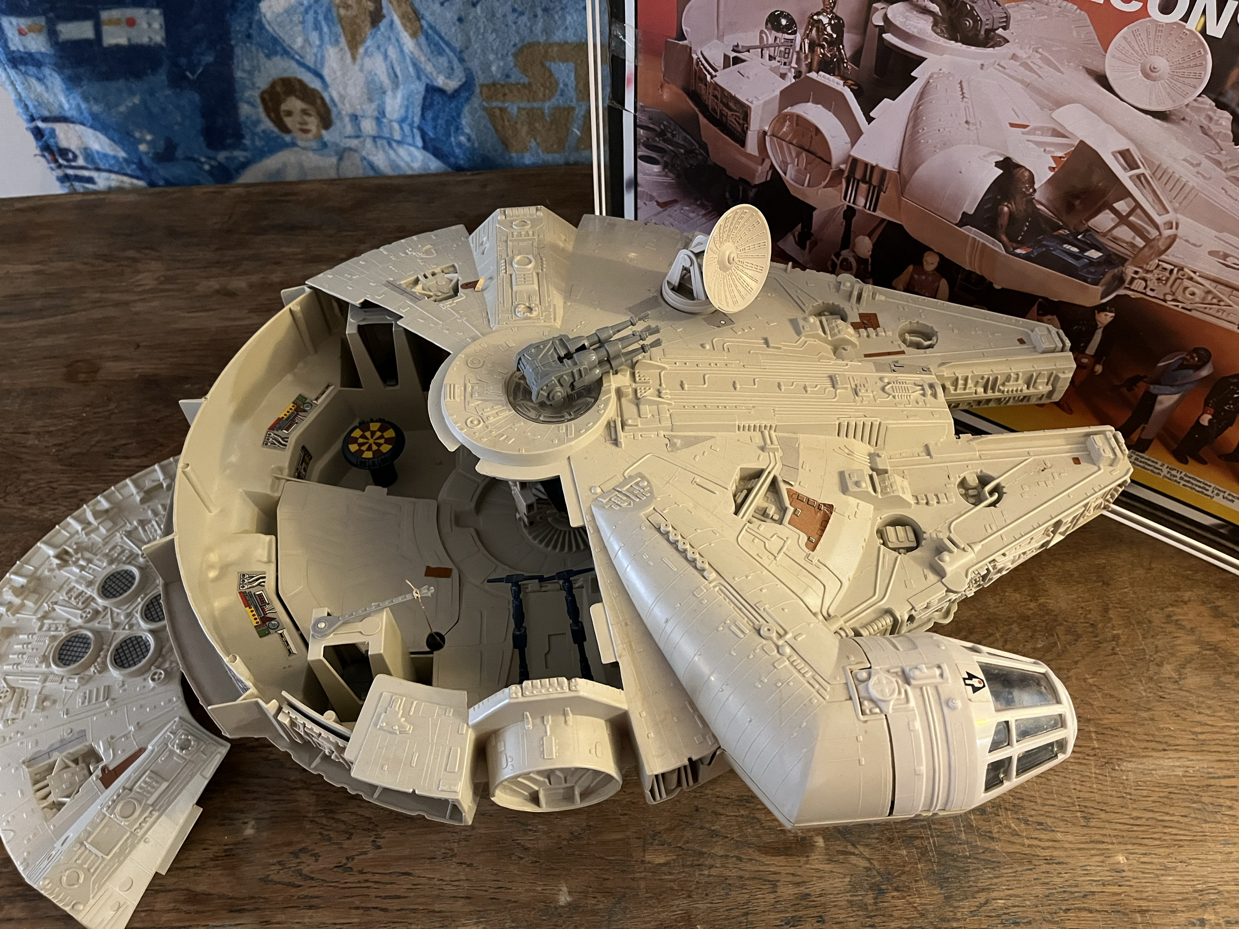 Close-up of a partially assembled Millennium Falcon model from Star Wars, resting on a wooden surface. Background features a poster and a cloth with a patriotic star pattern.