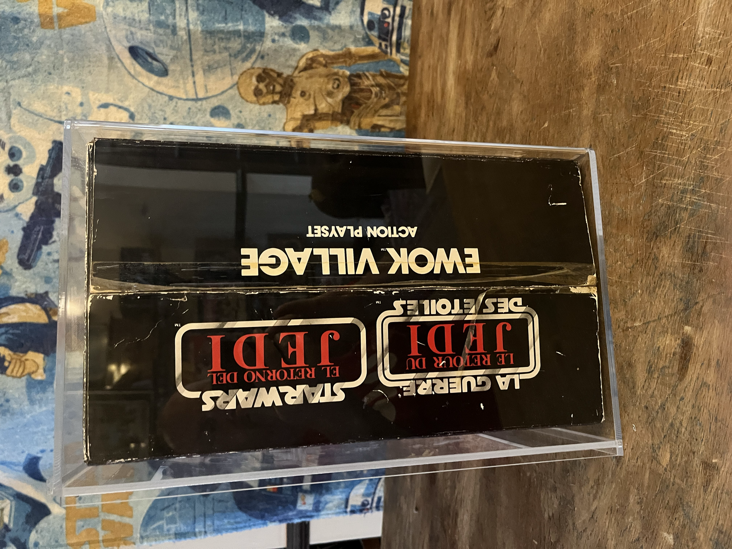 A clear plastic display case on a wooden surface contains a set of black trading cards with red and white text. In the background, there is a blue mural with vintage airplane illustrations and a wooden sculpture of a bird or animal.